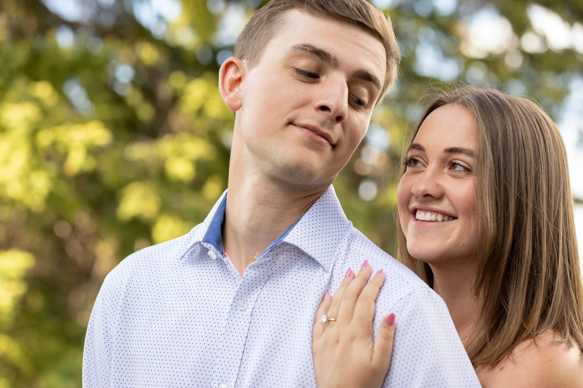 A young man and woman outdoors, close together, smiling and gazing at each other, with a background of green trees and sunlight.
