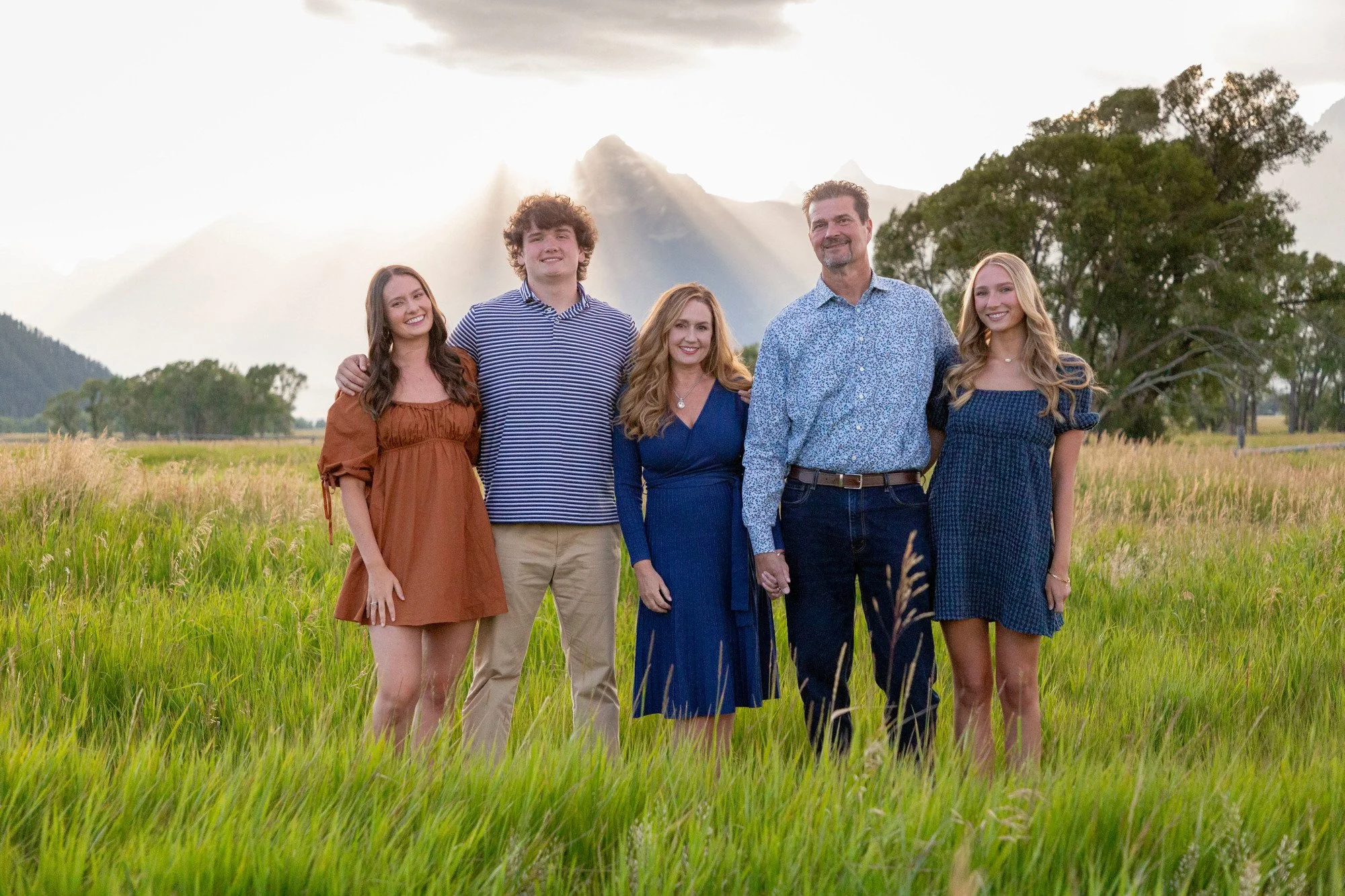 A family of five standing together in a field of tall grass with mountains and trees in the background during sunset. The family consists of three women, one man, and one young man, all smiling and dressed in semi-formal attire.
