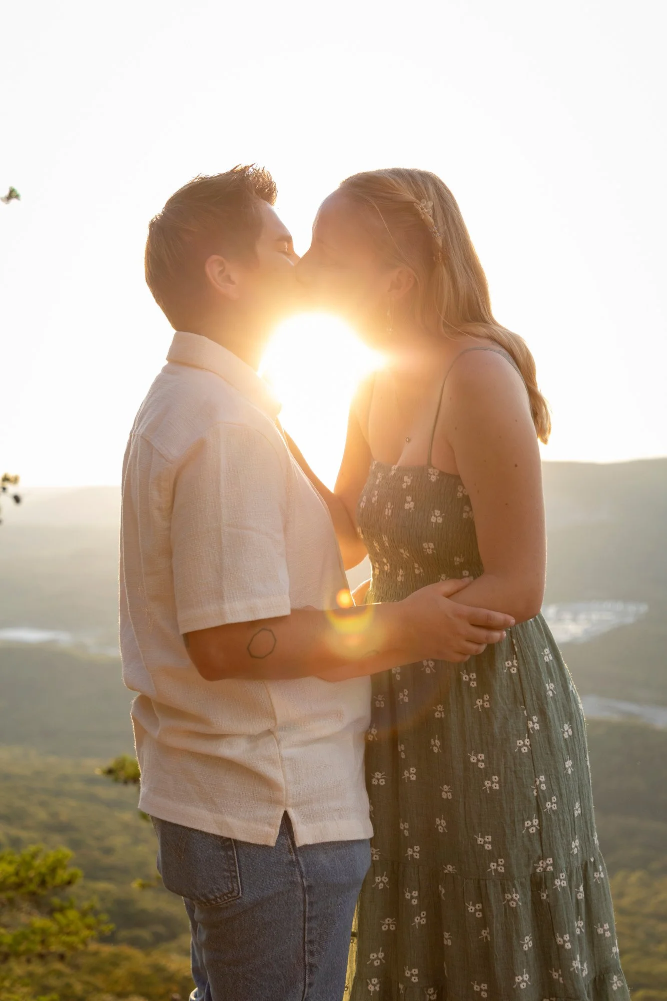 A couple sharing a kiss outdoors at sunset, with the sun glowing between them, on a hillside with lush greenery.