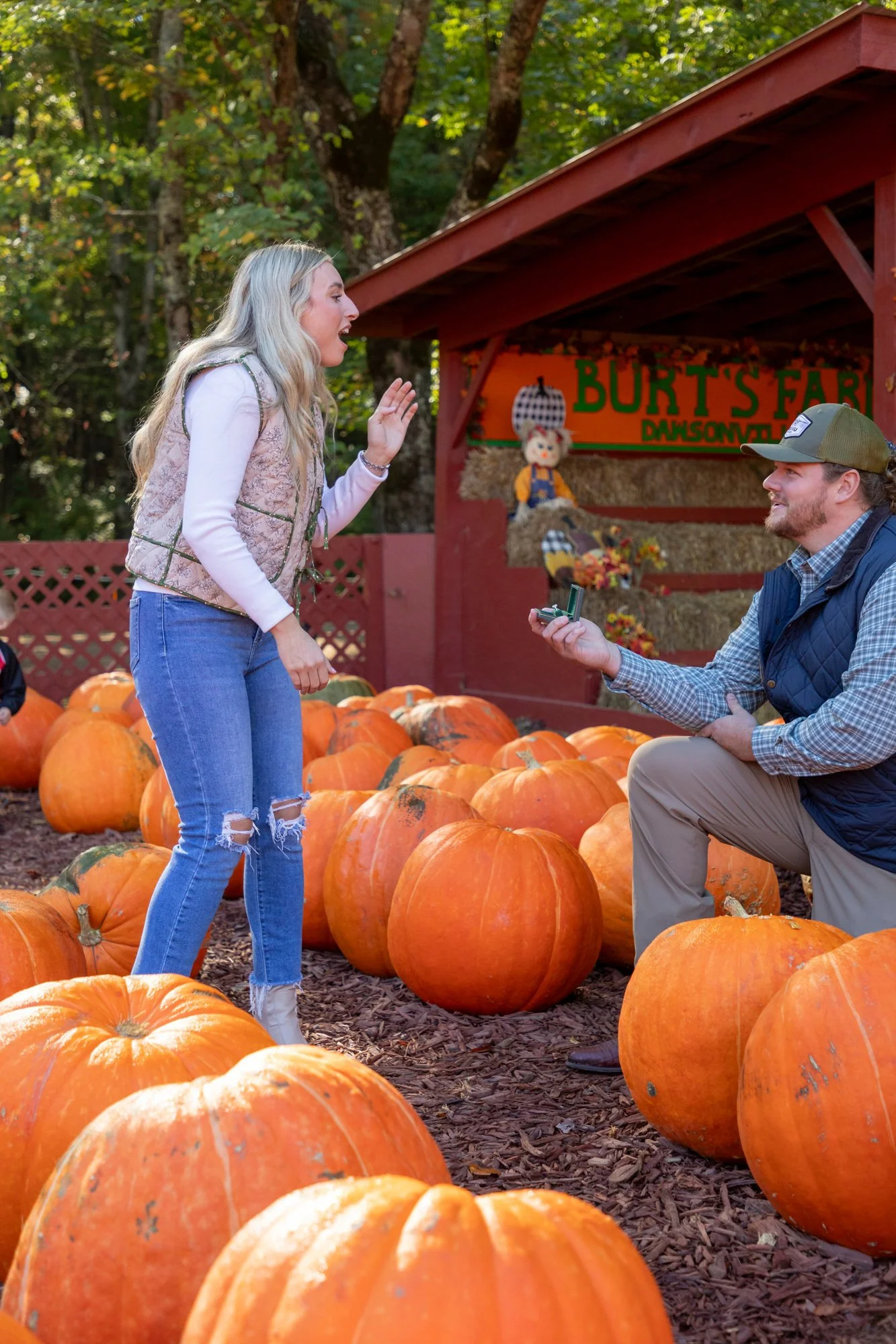 A woman with long blond hair, wearing a pink vest and ripped jeans, is talking to a man seated on a pumpkin at a pumpkin patch. The man is wearing a green cap, a blue vest, and khaki pants, and is holding a small item. Pumpkins are on the ground arou