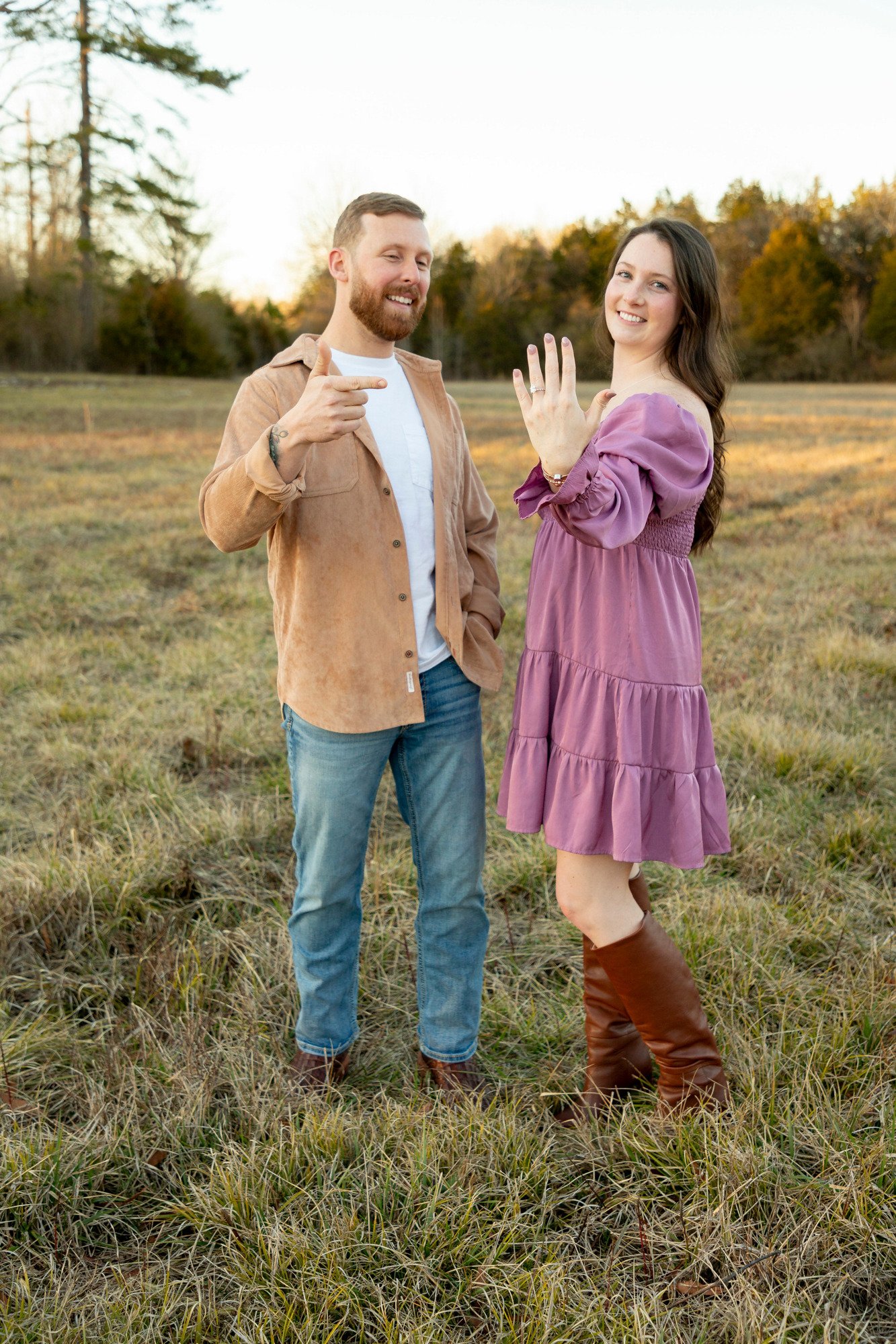 A man and woman standing in a field outdoors during fall, smiling, with the woman displaying an engagement ring.