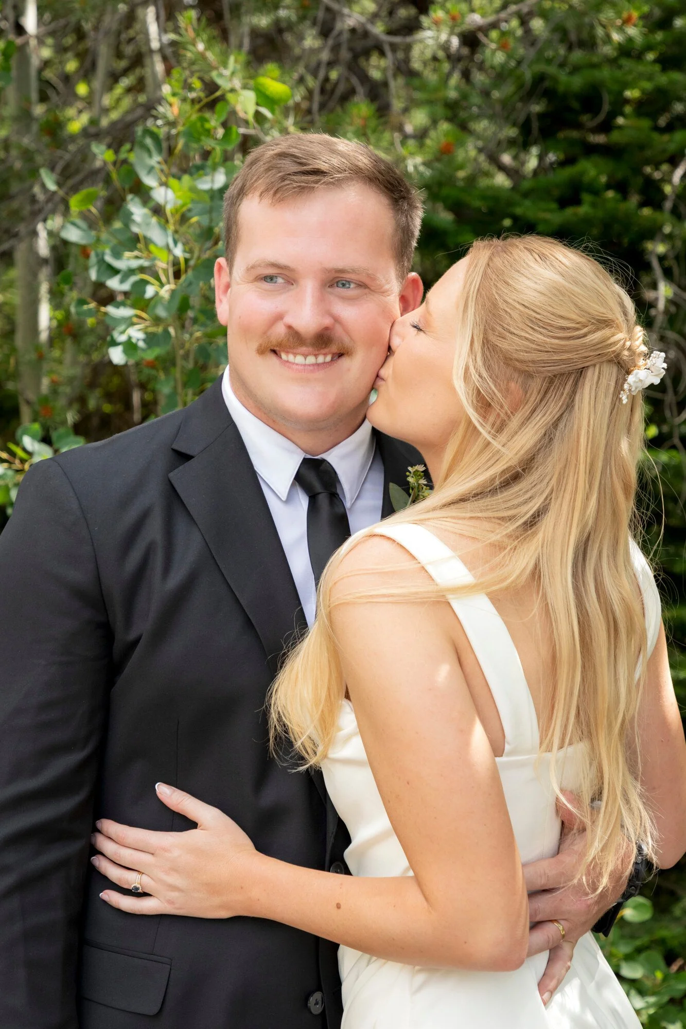 A newlywed couple in wedding attire, with the bride kissing the groom on the cheek outdoors with green trees in the background.