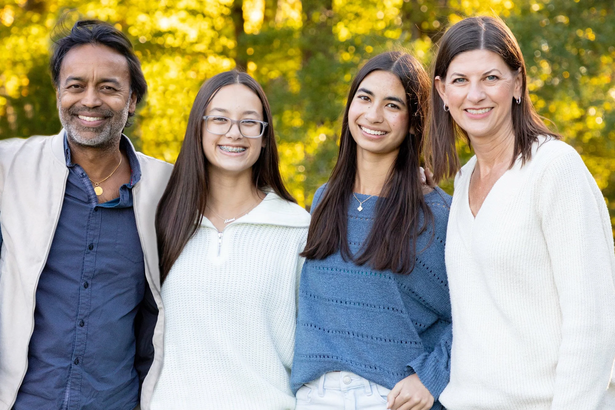 A diverse group of four people, two adults and two teenagers, standing outdoors in a park with green and yellow autumn leaves. They are smiling and appear to be enjoying a pleasant day.