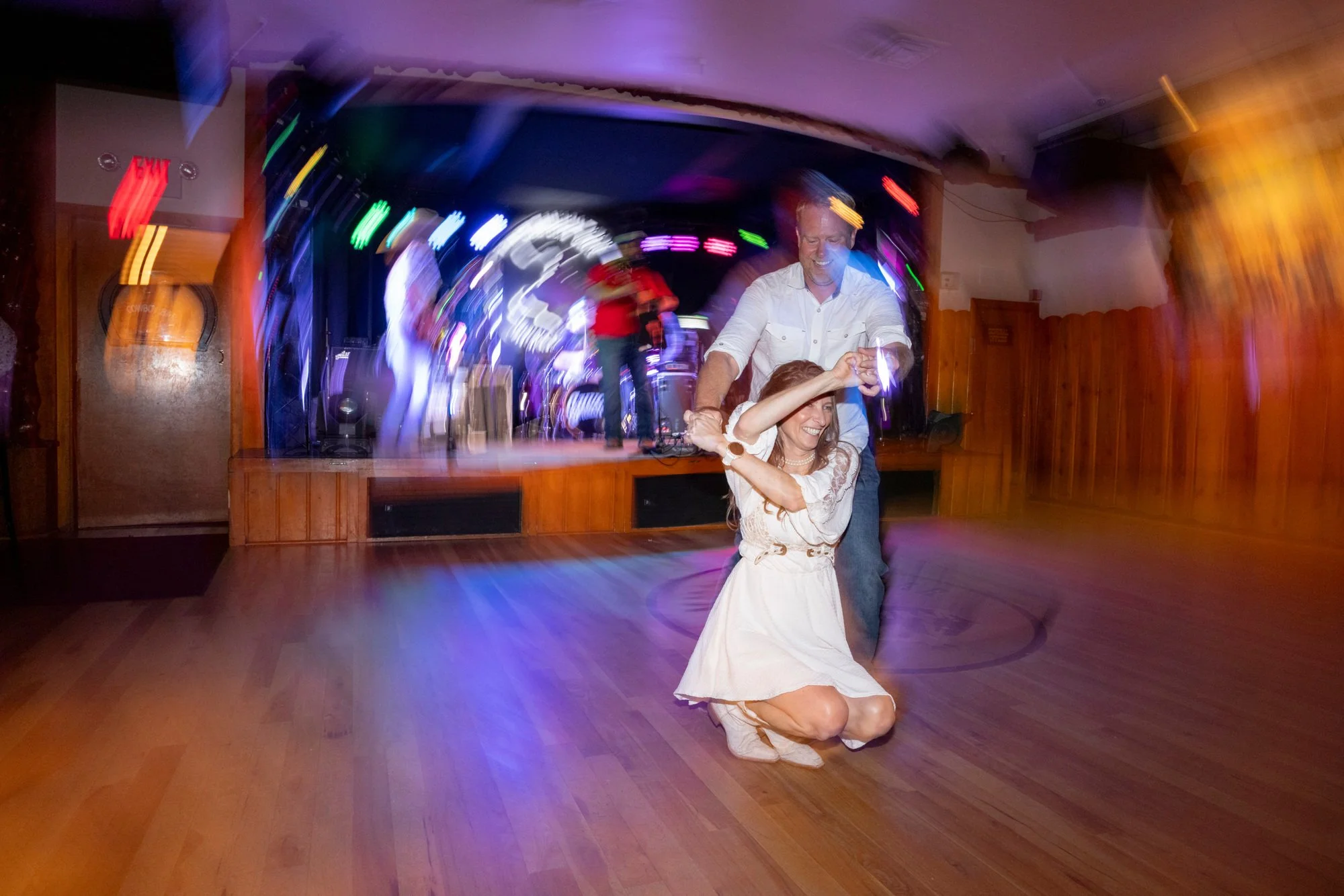 A couple dancing at a party or wedding reception with colorful lights and a live band playing on stage in the background.