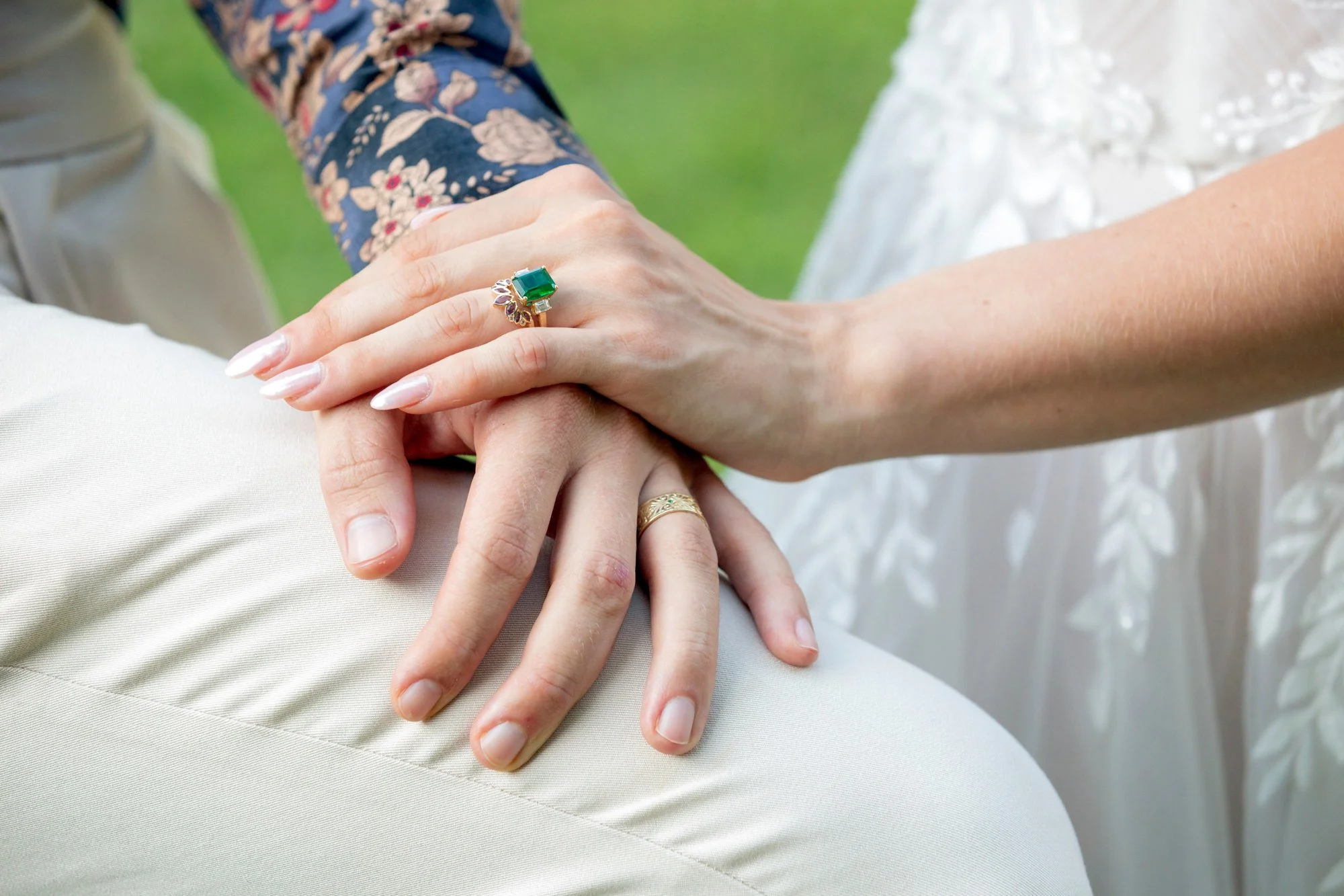 Close-up of two hands holding each other, one with an emerald ring, another with a gold ring, against green grass with a person in floral sleeve and a white dress visible in the background.