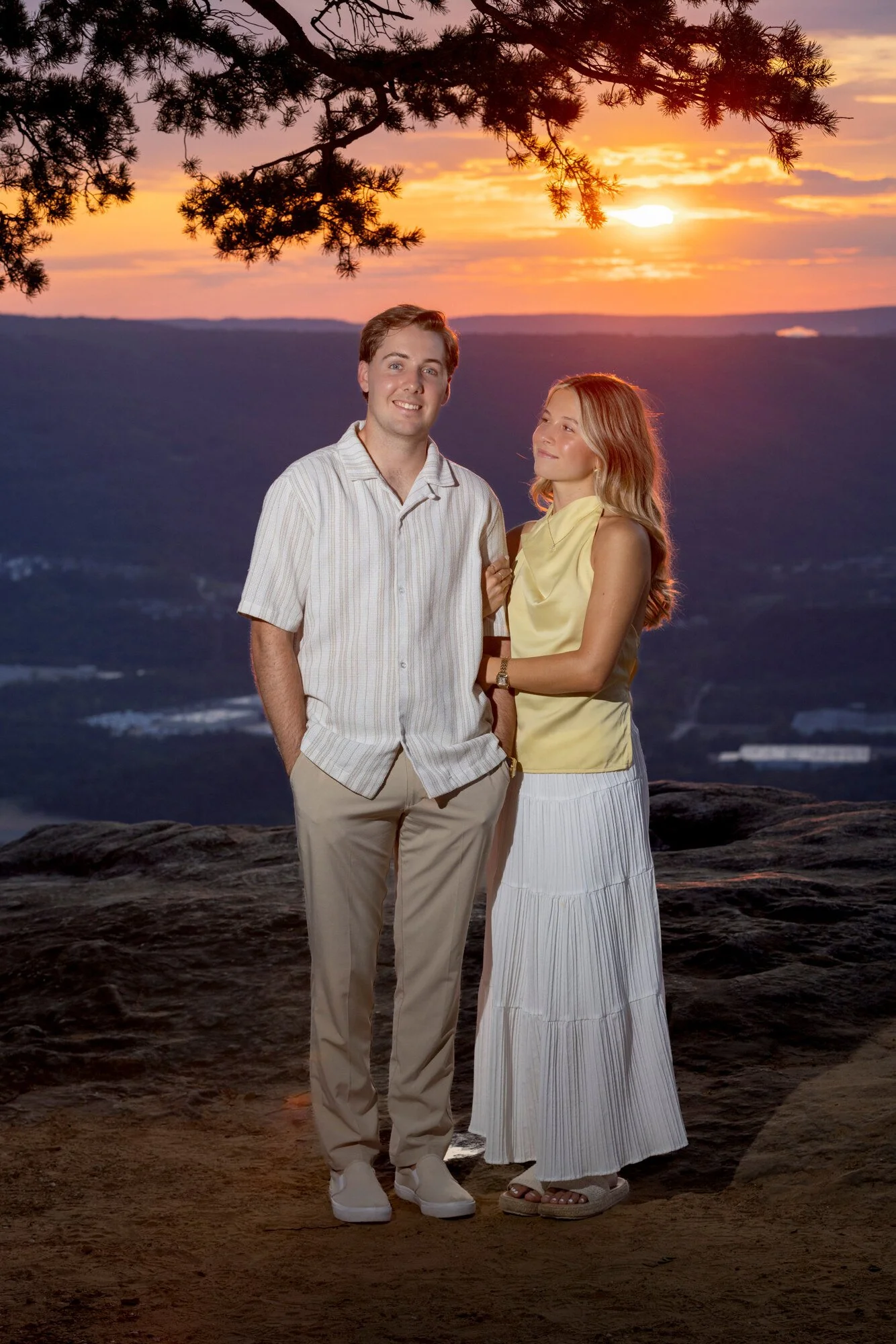 A young couple standing outdoors during sunset, with a scenic mountain landscape in the background, trees above, and rocky ground beneath them.
