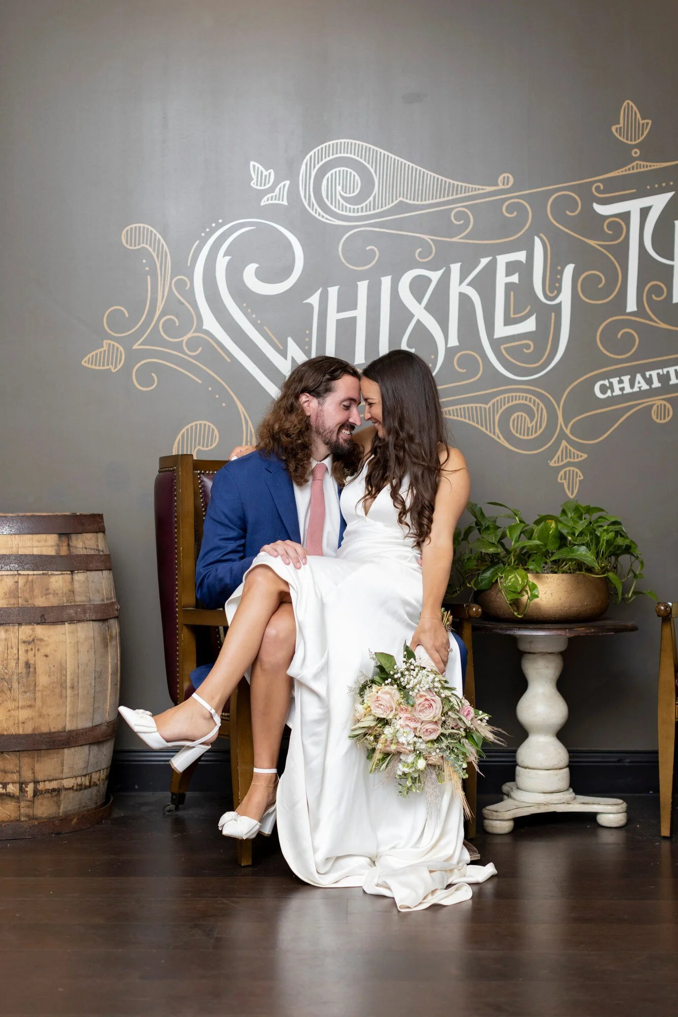 A couple dressed in wedding attire poses intimately in a rustic setting with props like a barrel and a potted plant, featuring a gray wall decorated with white lettering and swirls.