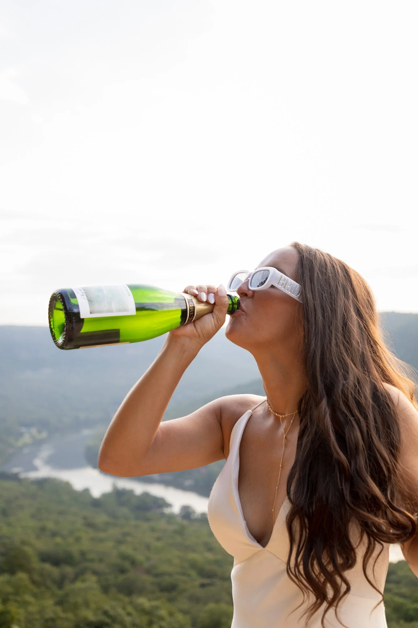 A woman with long brown hair and white sunglasses drinking from a green champagne bottle outdoors against a backdrop of green hills and a cloudy sky.
