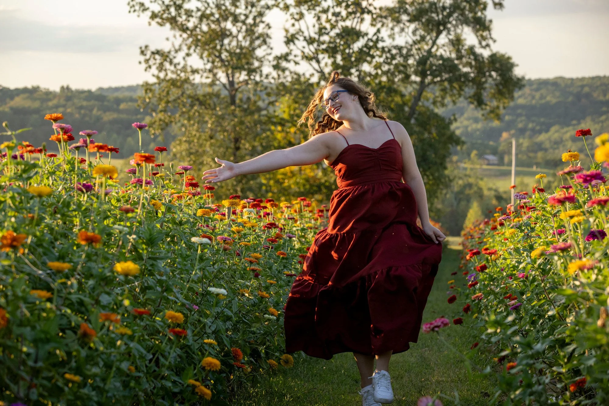 A woman in a maroon dress with spaghetti straps and glasses is running through a flower field. She is smiling and extending her right arm. The field is filled with colorful flowers and there are trees and hills in the background.