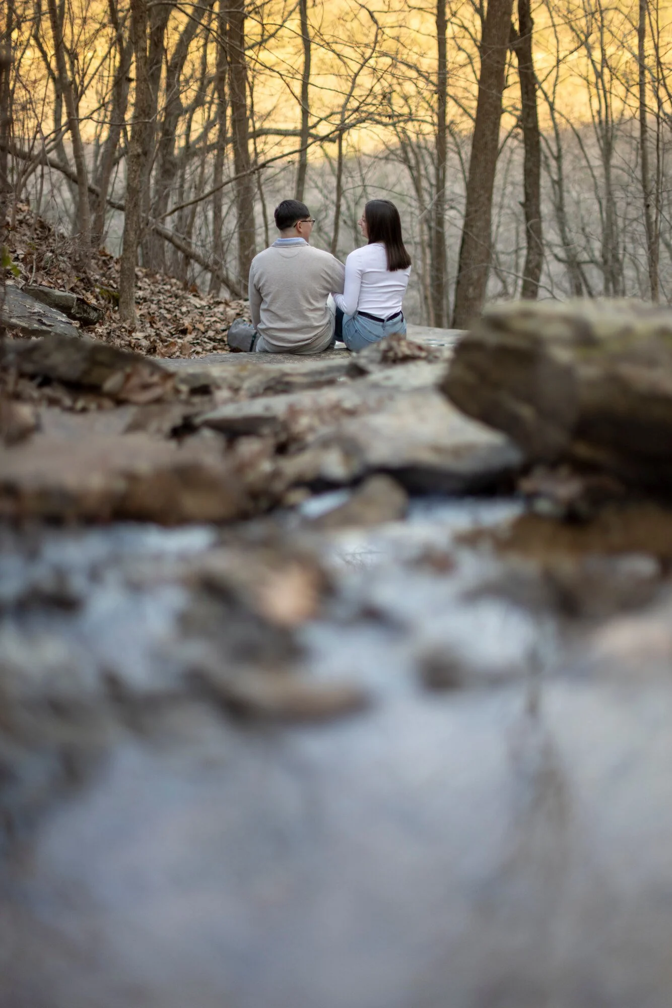 A man and woman sit on a log in a wooded area during sunset, facing each other and engaged in conversation.