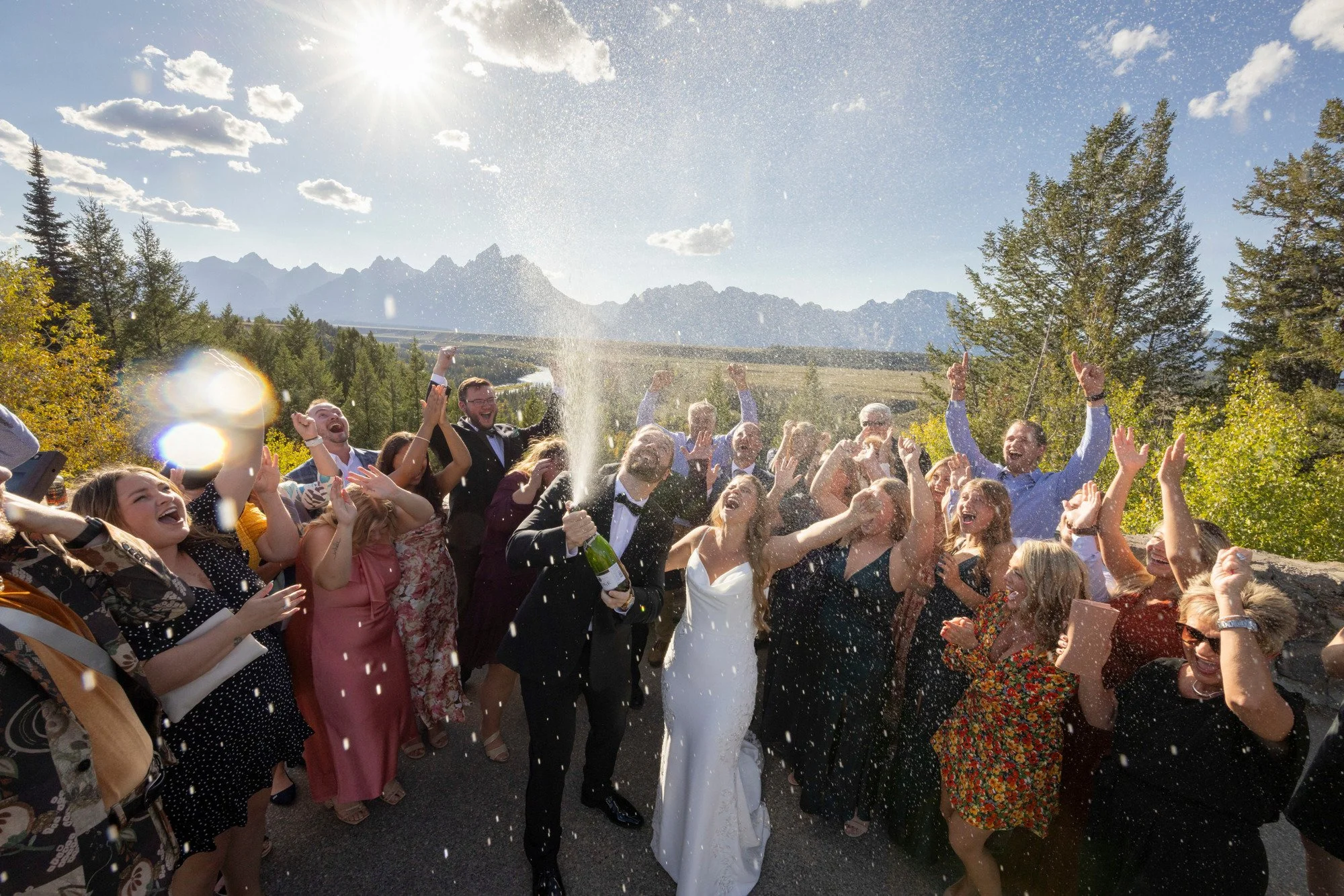 A group of people at a celebration outdoors, with some throwing champagne and celebrating under a bright, sunny sky with mountains and trees in the background.