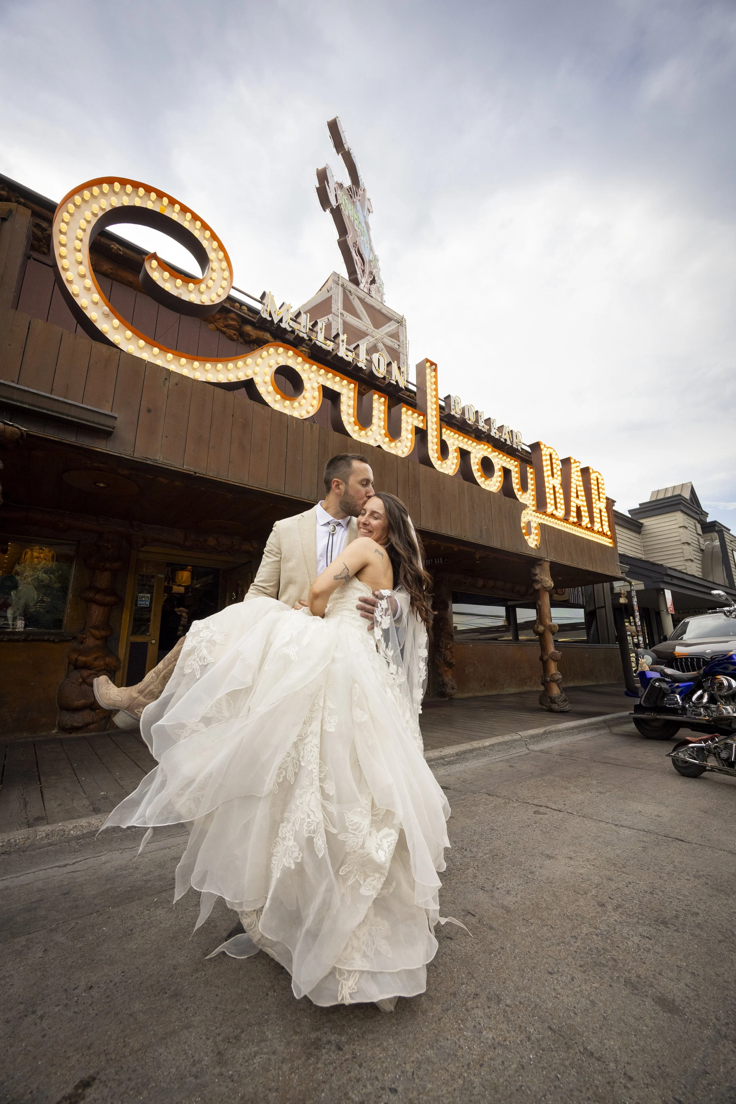 A newlywed couple sharing a tender moment outside a rustic bar, with the bride wearing a long white wedding dress and the groom in a light-colored suit, in front of a wooden building with a vintage sign reading 'Honolulu Bar'.