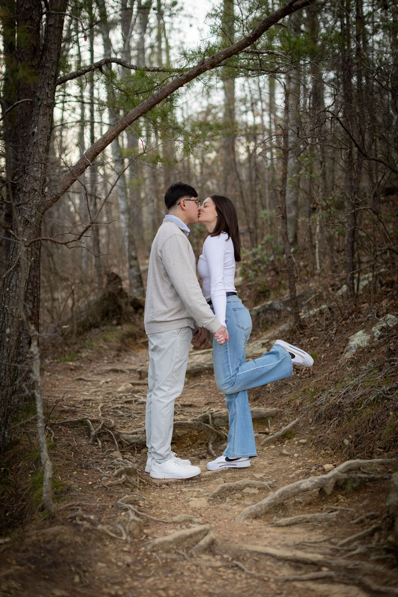 A young couple kissing in a forest, with the woman kneeling on the ground and holding the man's hands, surrounded by tall trees and a dirt path.