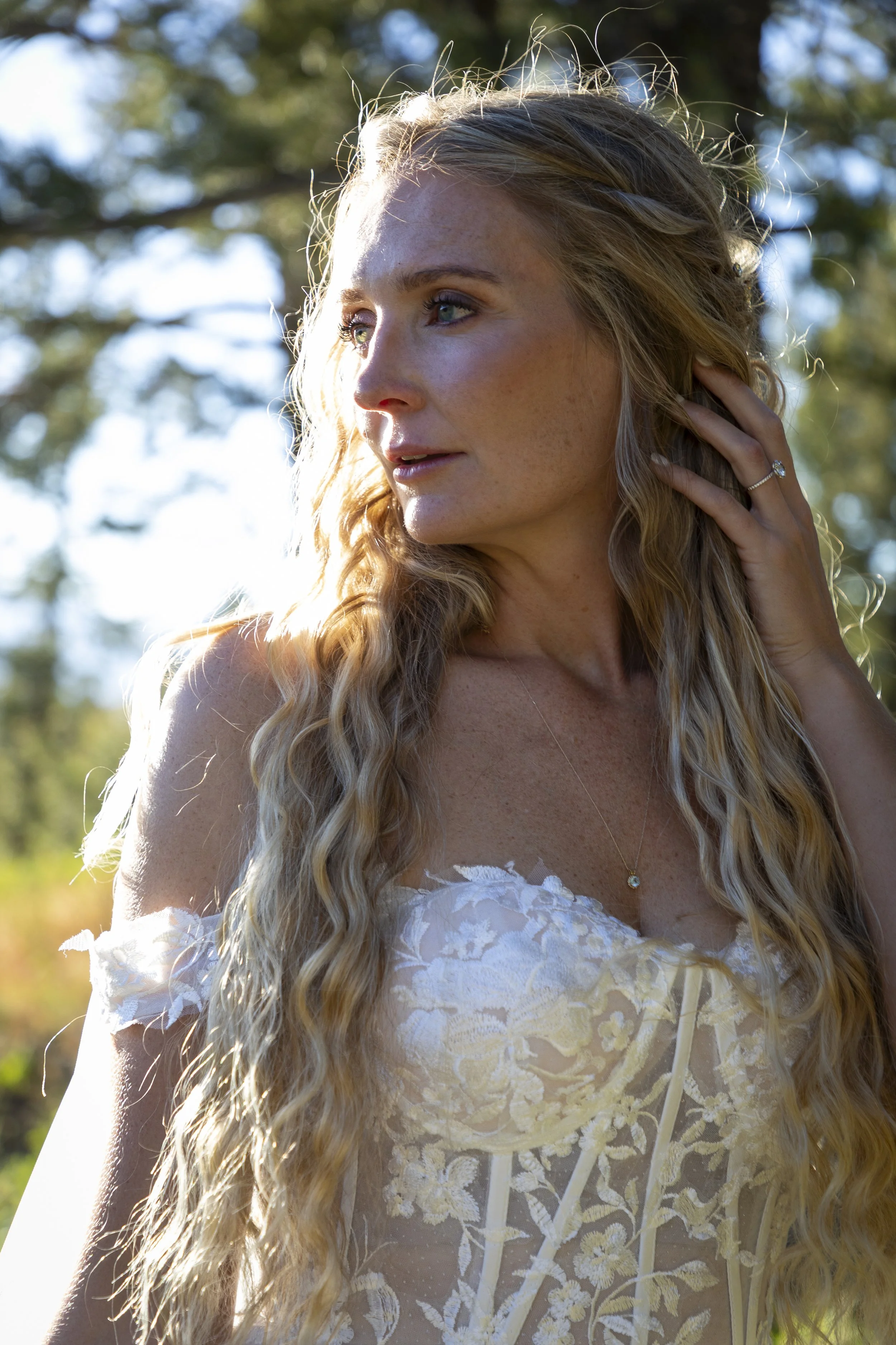 A woman with long, wavy, blonde hair, wearing a white lace dress and a delicate gold necklace, stands outdoors with sunlight illuminating her face and hair, surrounded by trees.