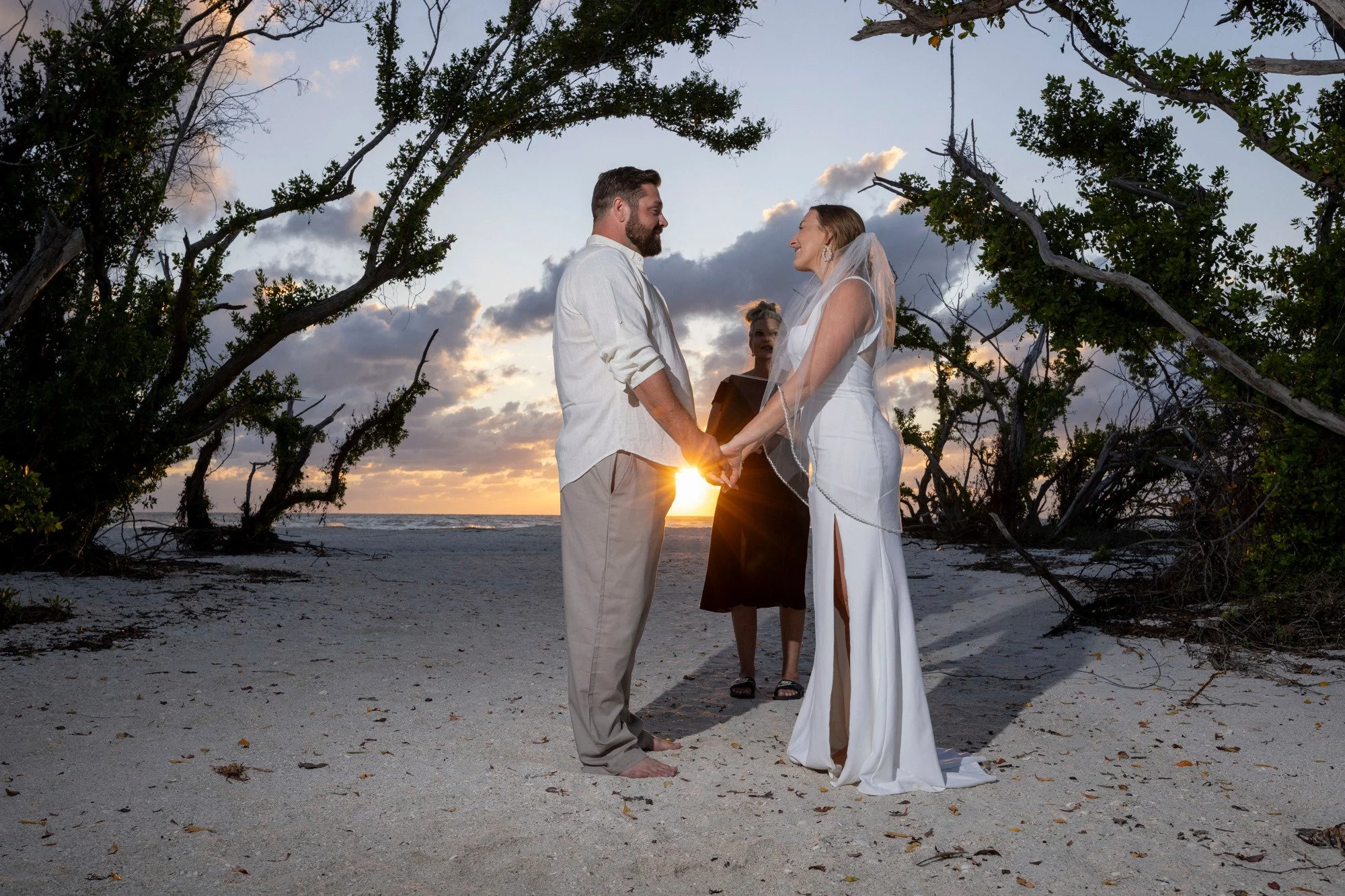 A couple getting married on the beach at sunset, holding hands and facing each other, with an officiant standing behind them and trees framing the scene.
