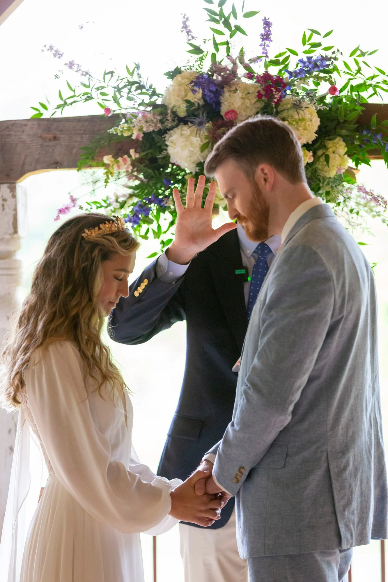 A couple holding hands during a wedding ceremony, with an officiant standing behind them, and a floral arrangement with white and purple flowers in the background.