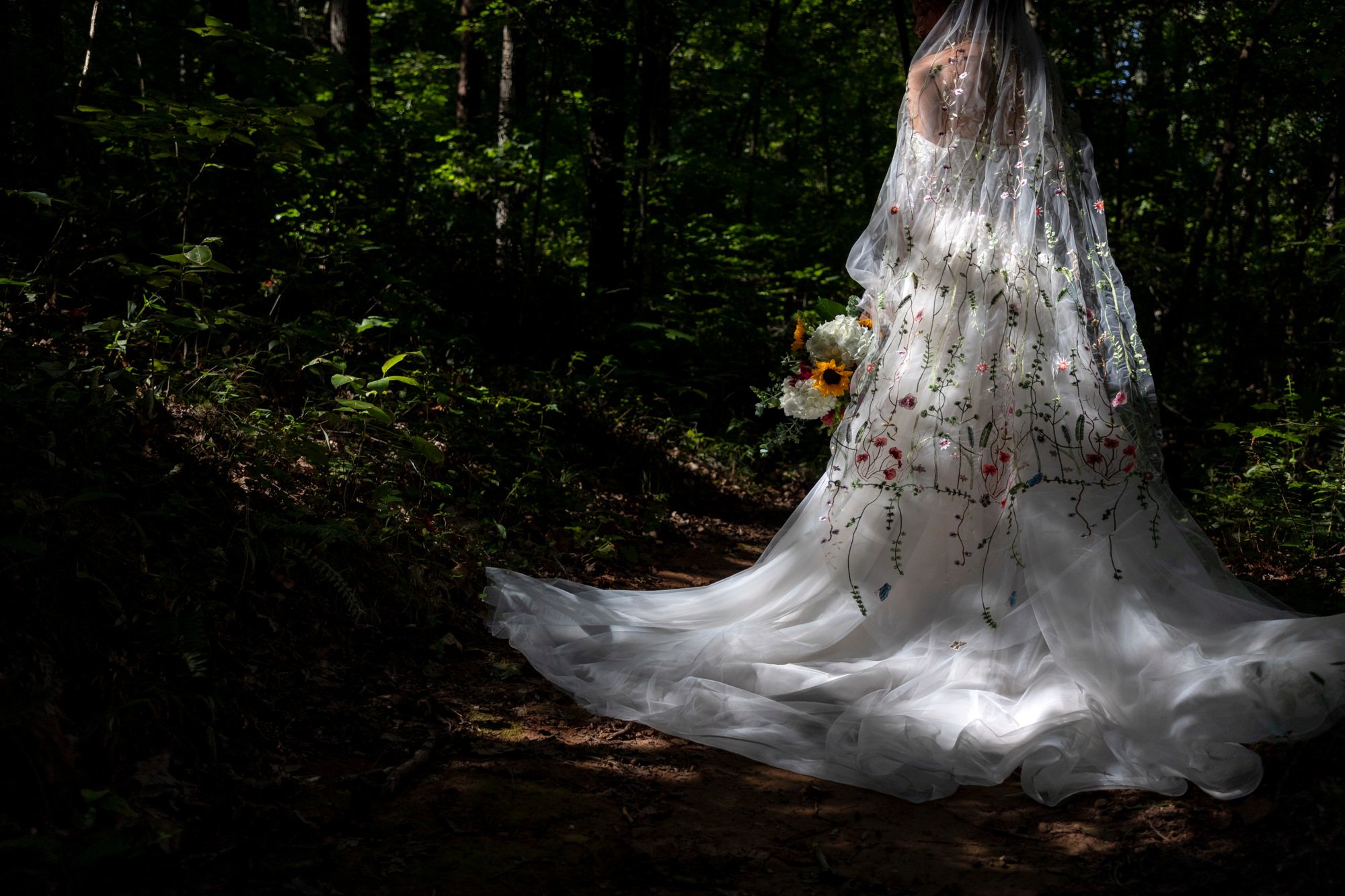 A bride wearing a white wedding dress with floral embroidery, standing in a shaded forest, holding a bouquet of flowers.
