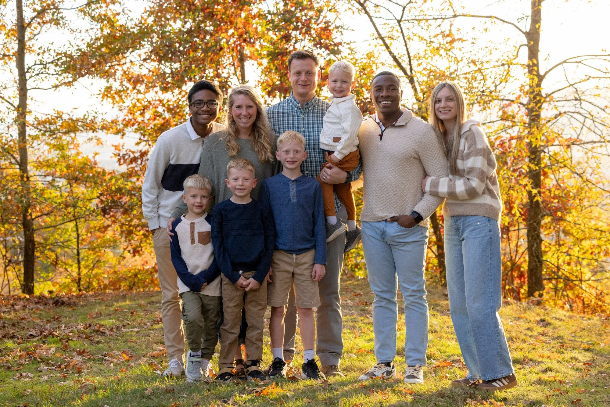 Group of ten people, including children and adults, standing outdoors in fall setting with trees and colorful autumn leaves in background, smiling at the camera.