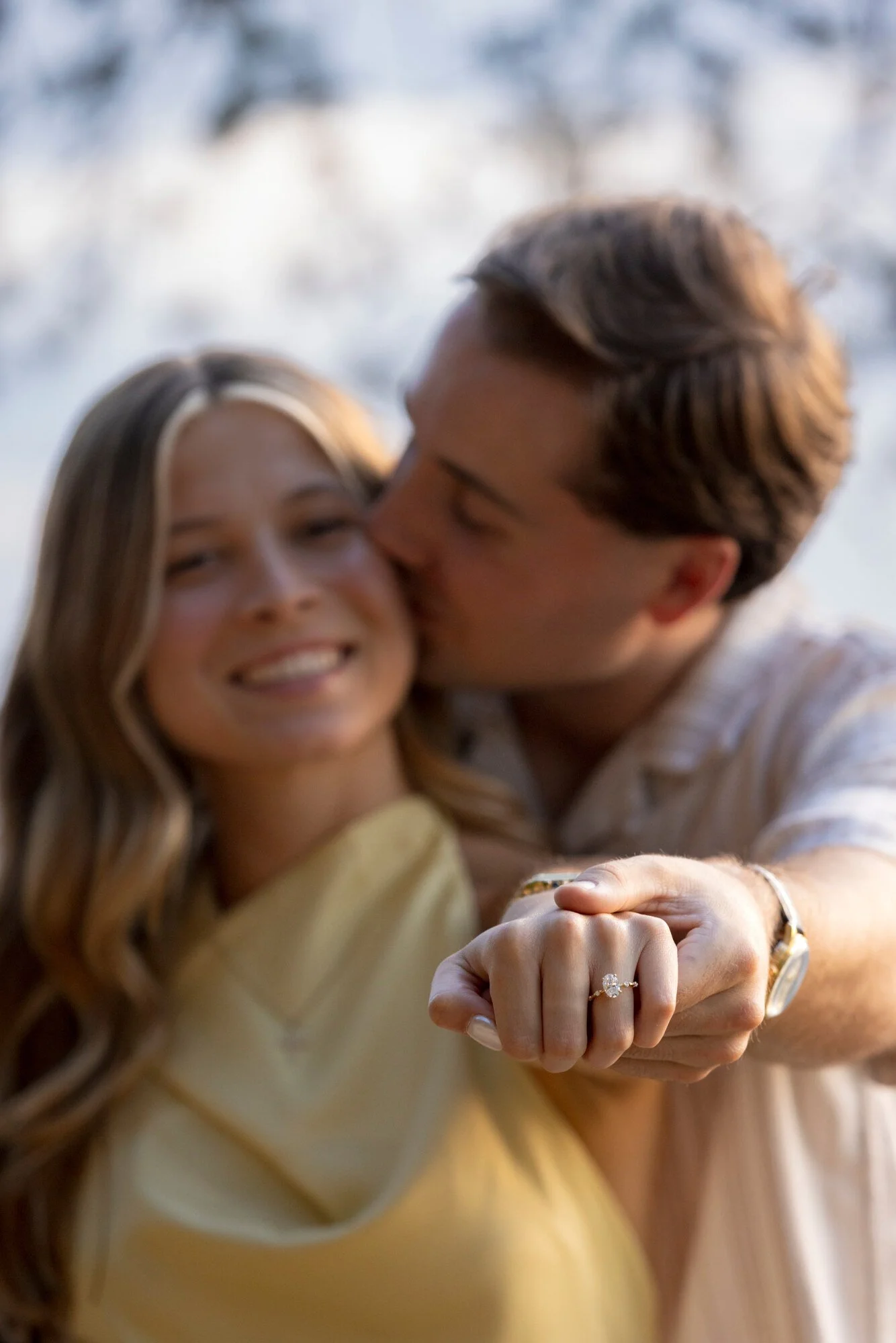 A young couple takes a selfie, with the woman smiling and showing her engagement ring, while the man kisses her on the cheek. The focus is on their hands and rings, with a blurred outdoor background.