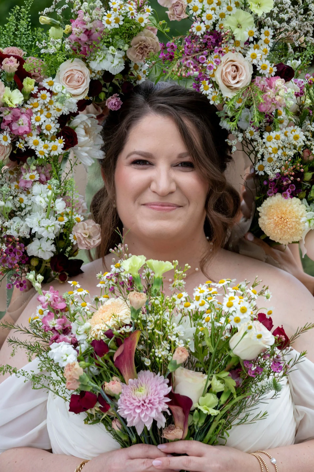 A woman wreathed with colorful flowers, holding a bouquet of mixed flowers, smiling softly.