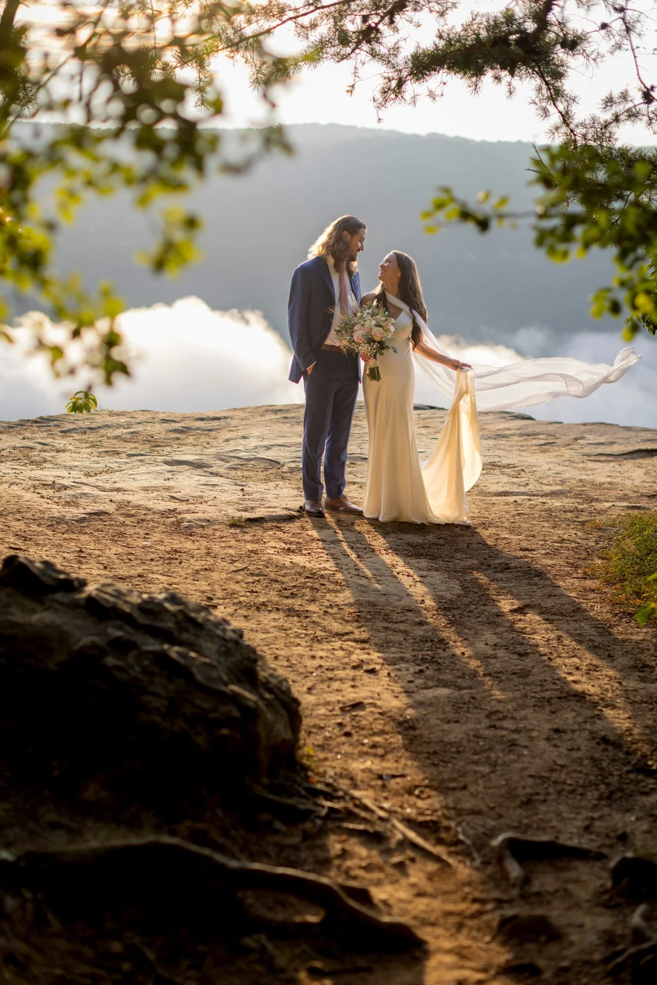 A bride and groom standing outdoors on a rocky surface, sharing a moment with the bride holding a bouquet of flowers, surrounded by trees and mountain scenery in the background during sunset.