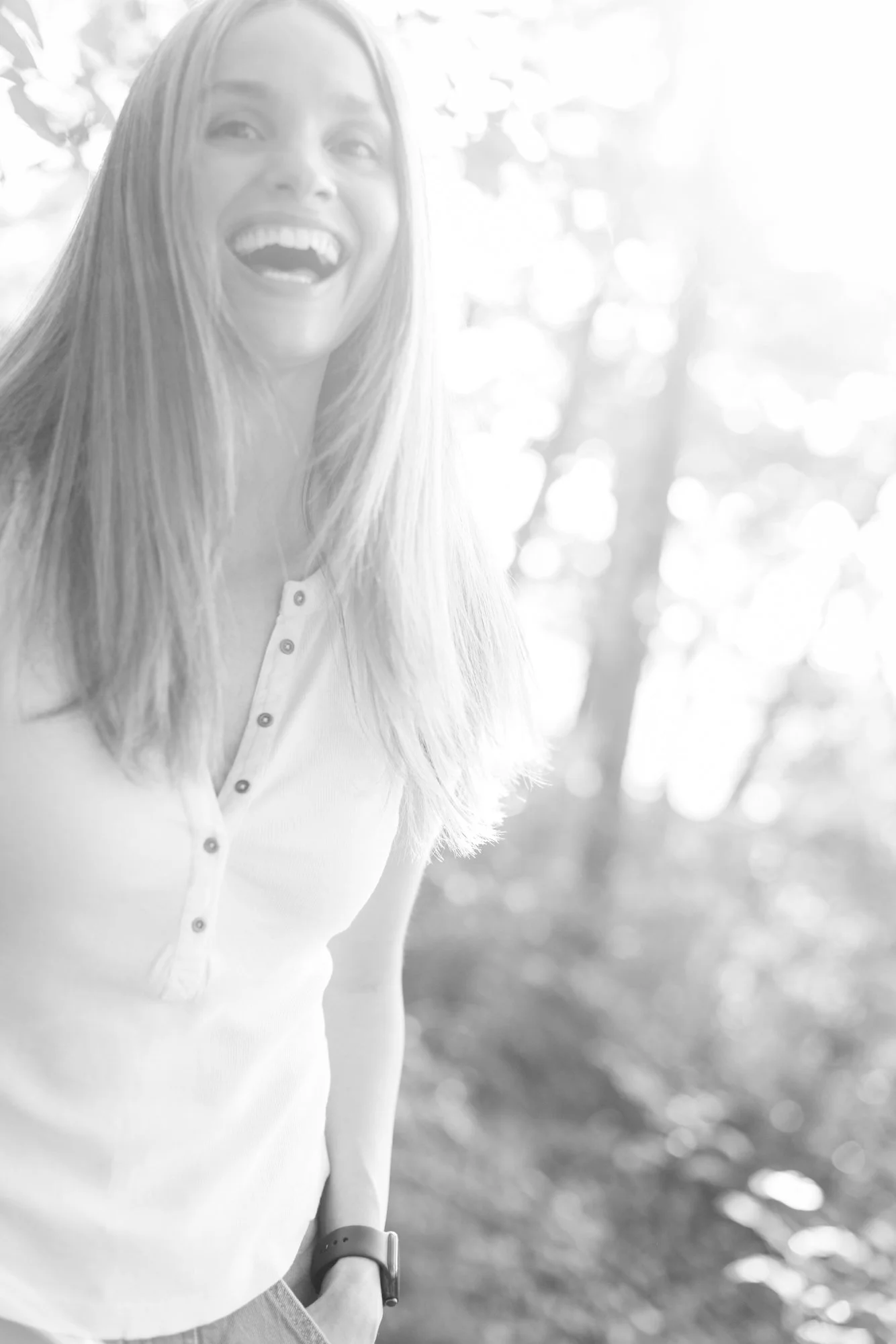 Black and white photo of a smiling woman outdoors, with her left arm bent at the elbow and wearing a watch, in a sunny background with bokeh lights and trees.