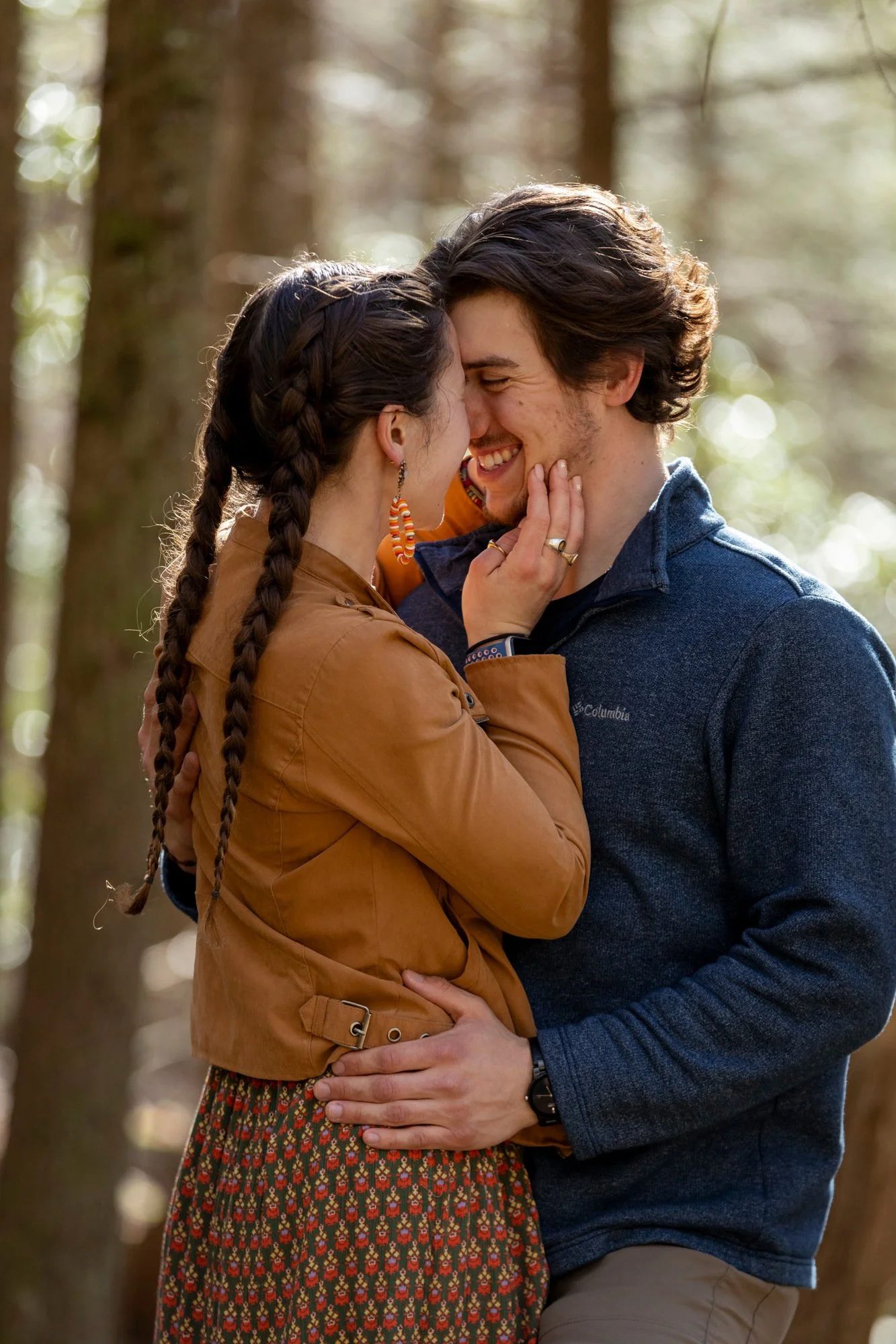 A couple smiling and touching foreheads in a forest during daytime, with sunlight filtering through trees.
