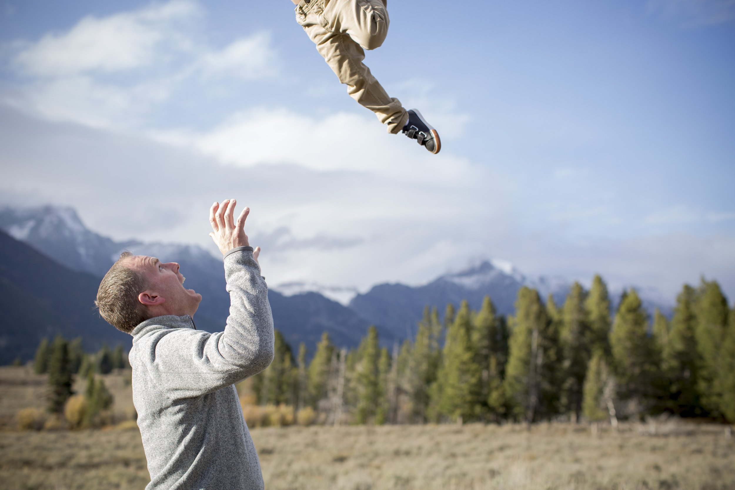 A young man in a gray hoodie catching a young child in a beige hoodie, both in an outdoor field with mountains, trees, and a blue sky with clouds in the background.