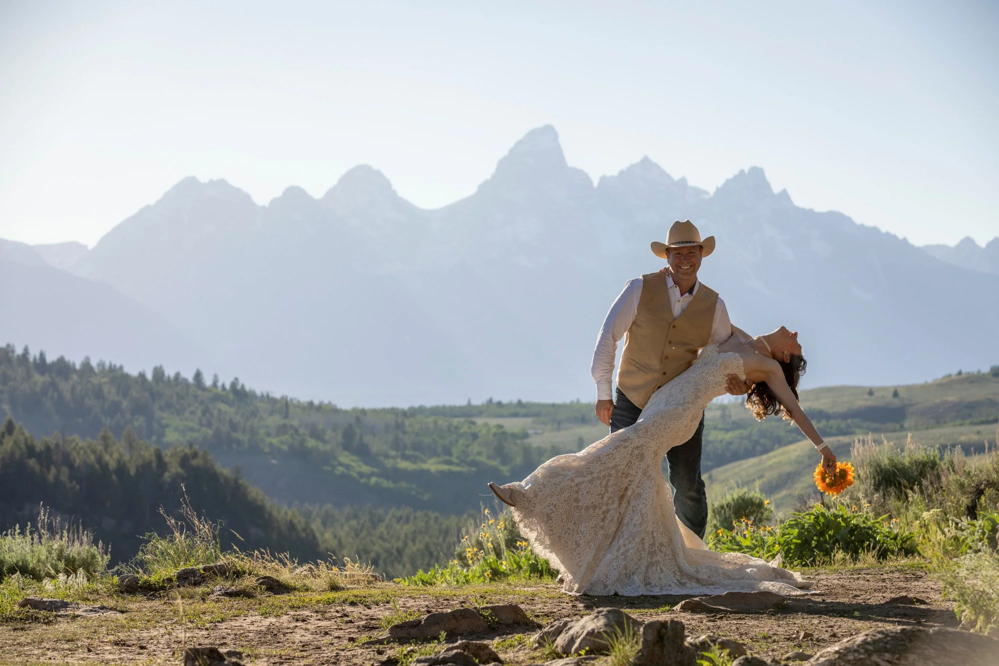 A man wearing a cowboy hat and vest and a woman in a wedding dress dancing outdoors in a field with mountains in the background, holding a sunflower bouquet.