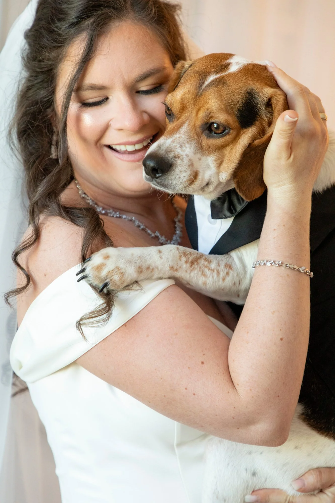 A woman with dark brown hair and a white dress hugging a brown and white dog wearing a tuxedo style collar.