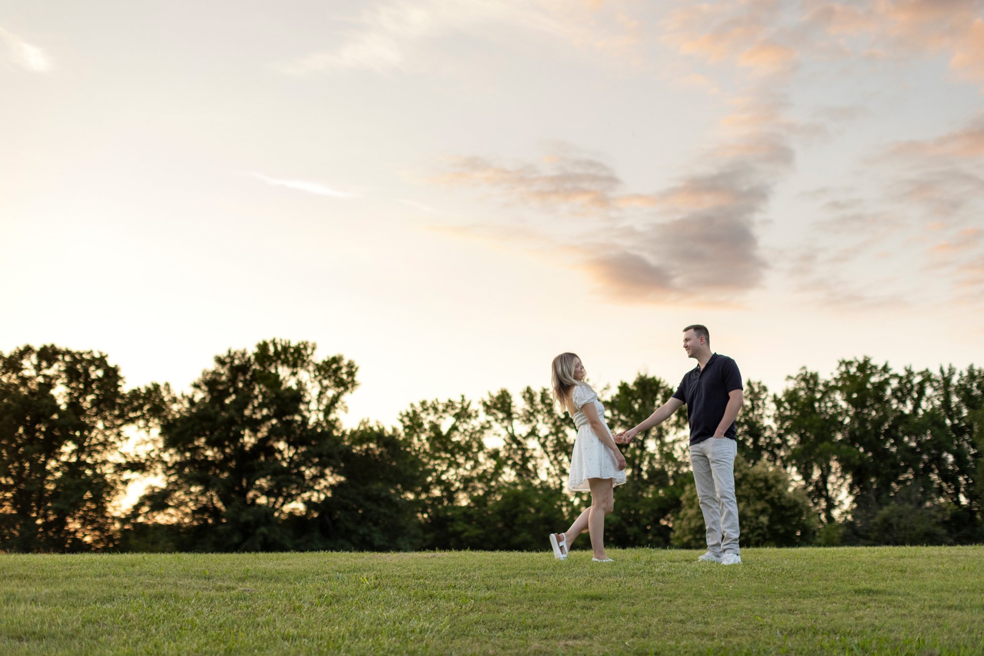 A young couple holding hands in a grassy field at sunset, with trees in the background and a partly cloudy sky.