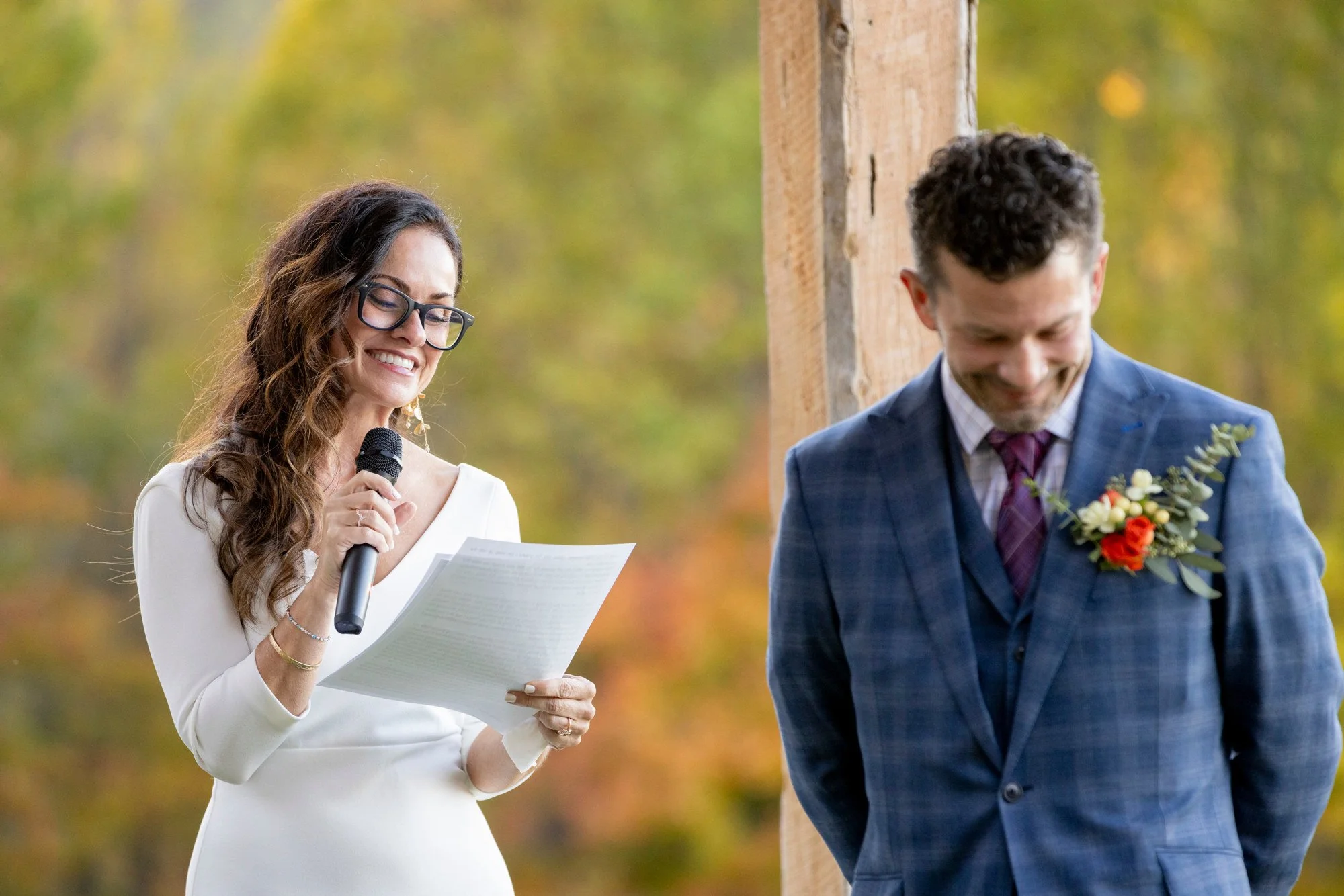 Woman with glasses reading from paper during outdoor wedding ceremony, man in suit with boutonniere, standing against wooden structure with autumn-colored trees in background.
