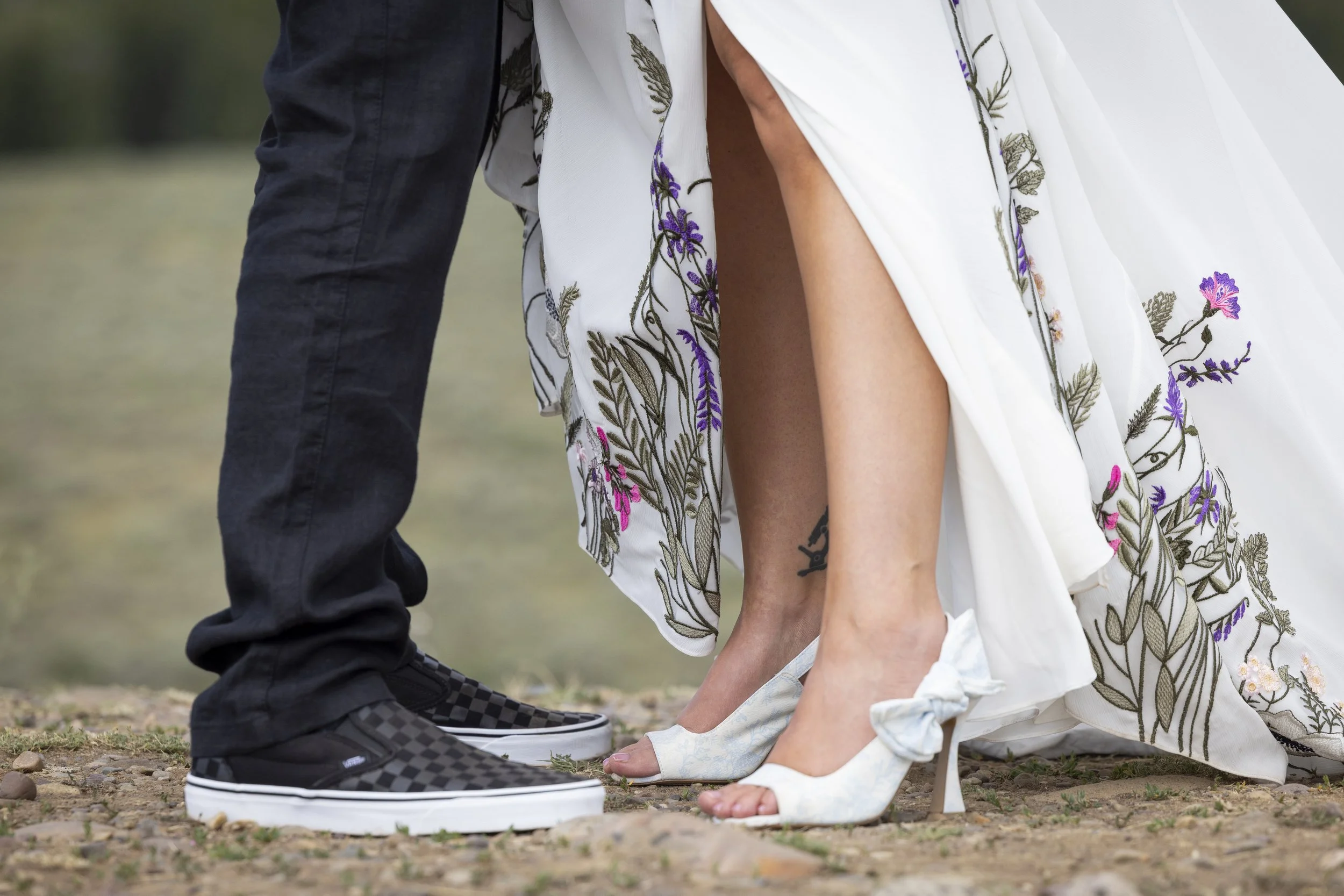 Close-up of a couple, with the woman dressed in a long white floral embroidered dress kneeling with her back slightly arched and the man in black pants and checkered shoes standing next to her on a grassy ground outdoors.