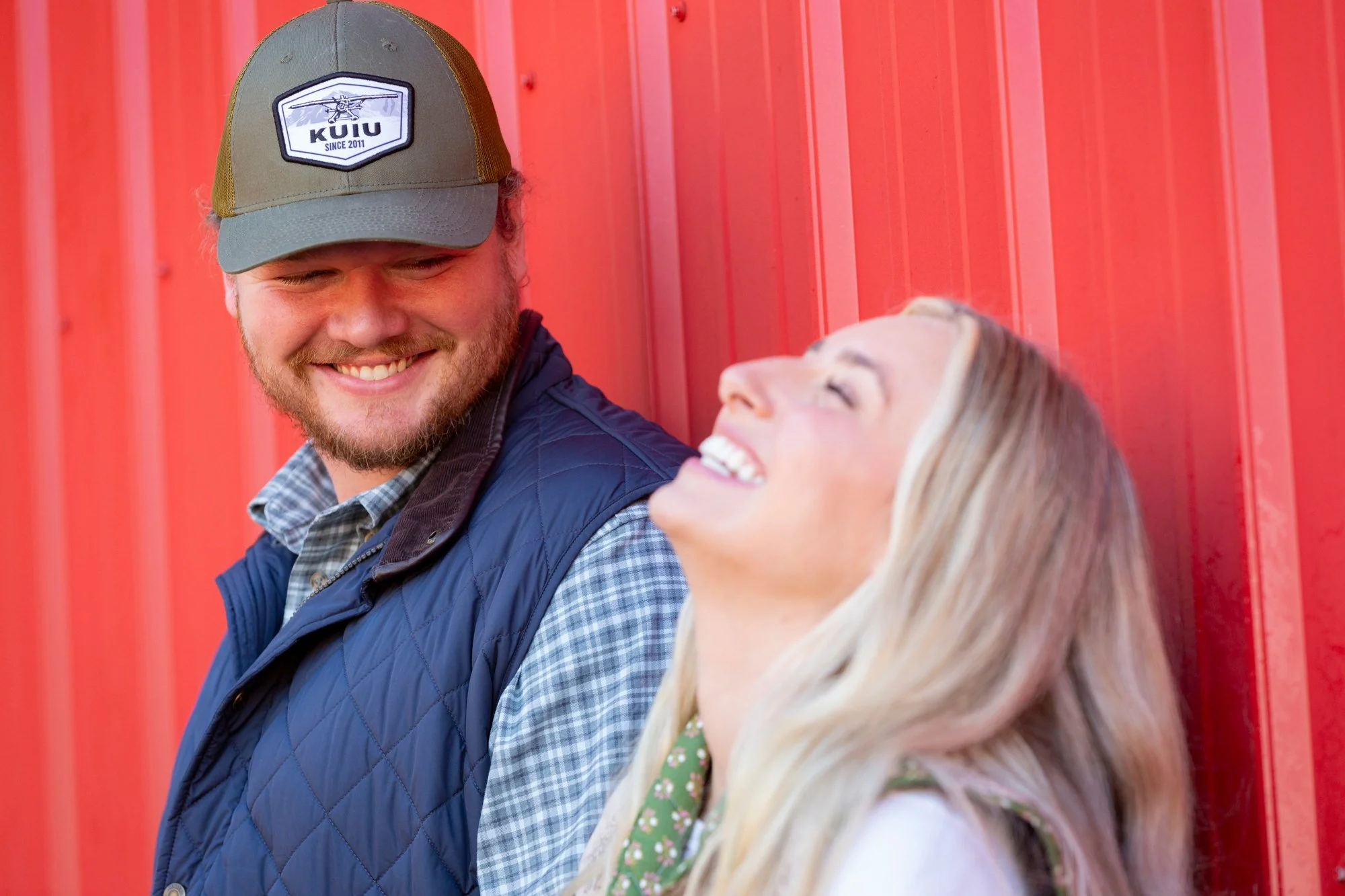 A young man and a young woman are smiling and looking at each other while leaning against a red metal wall.