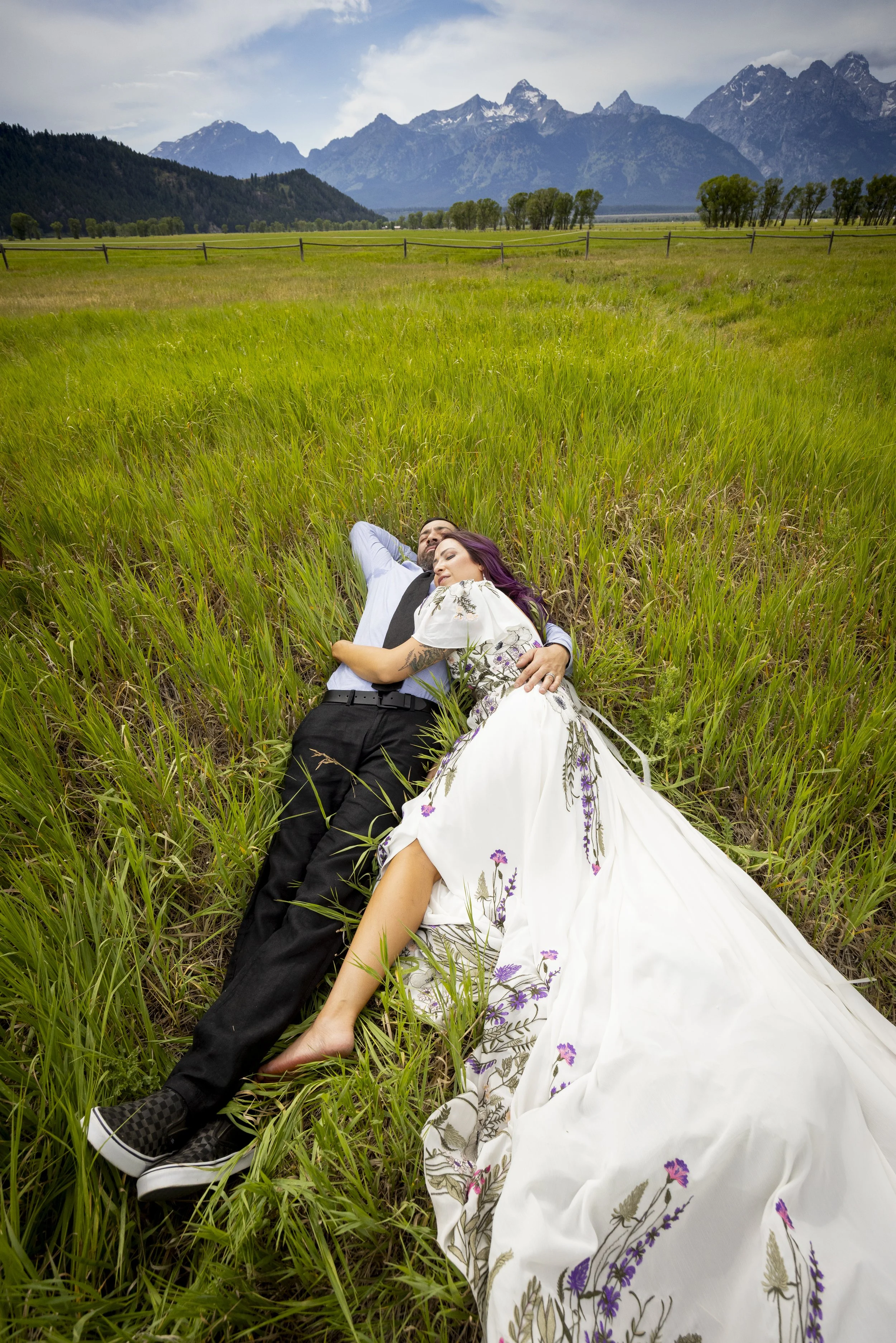 A man and woman lying together in a grassy field with mountains in the background, the man wearing a light blue shirt and black tie, and the woman in a white dress with floral embroidery, both with relaxed expressions.
