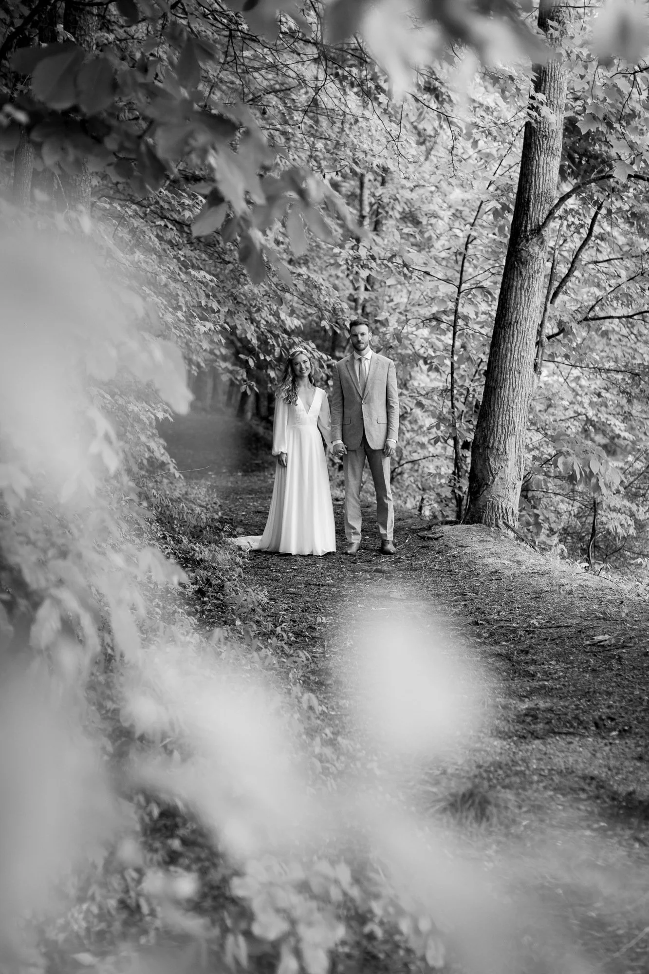 A black-and-white photo of a couple in wedding attire holding hands, standing on a wooded trail surrounded by trees and foliage.