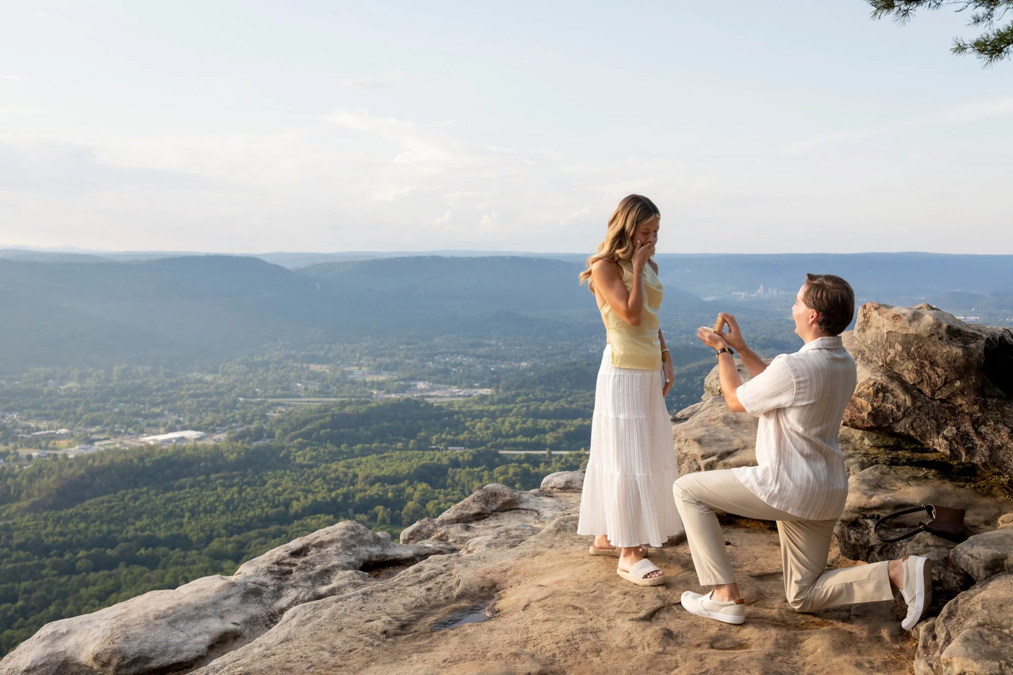A man proposing marriage to a woman on a rocky hill with a scenic view of a valley and distant mountains in the background, during a sunset or late afternoon. The woman looks surprised and emotional, while the man is kneeling and presenting a ring.