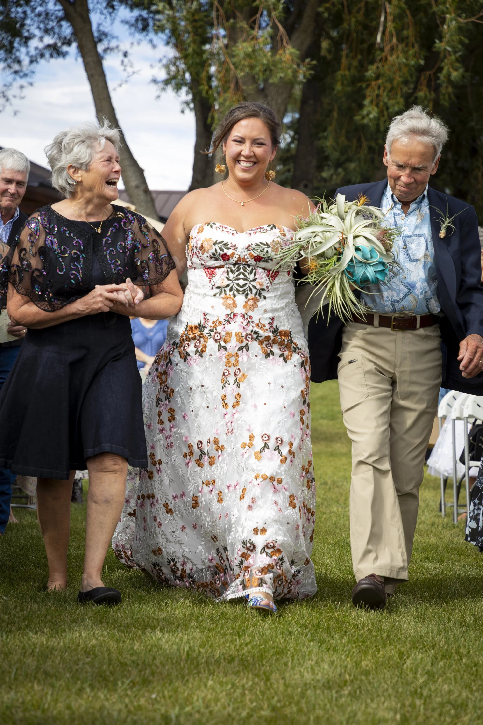A smiling bride holding a bouquet of flowers walking on a grassy lawn with family members during a wedding celebration outdoors.