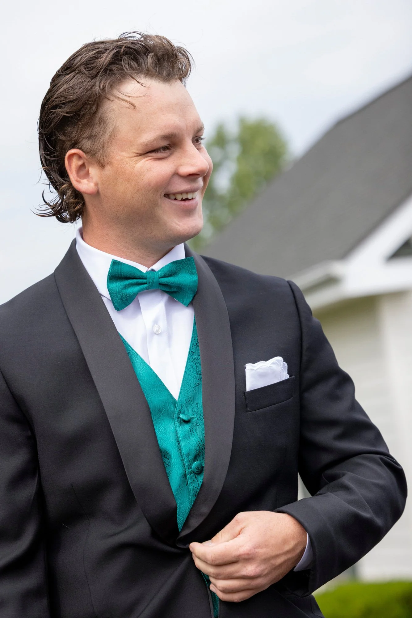 A man in a tuxedo with a teal bow tie and vest, smiling outdoors in front of a cloudy sky and some trees.