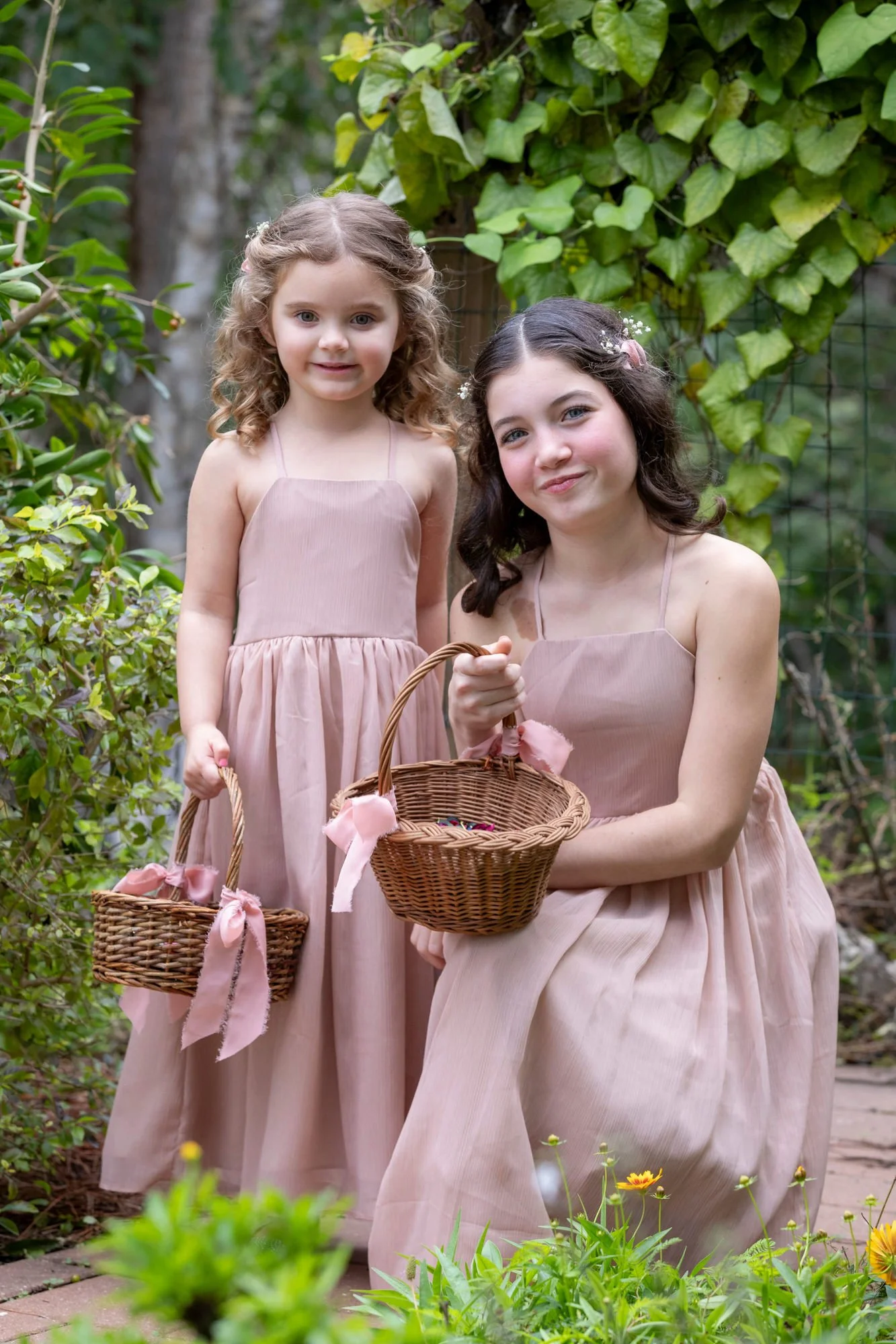Two young girls in matching pink dresses holding baskets and gathering flowers in a garden.