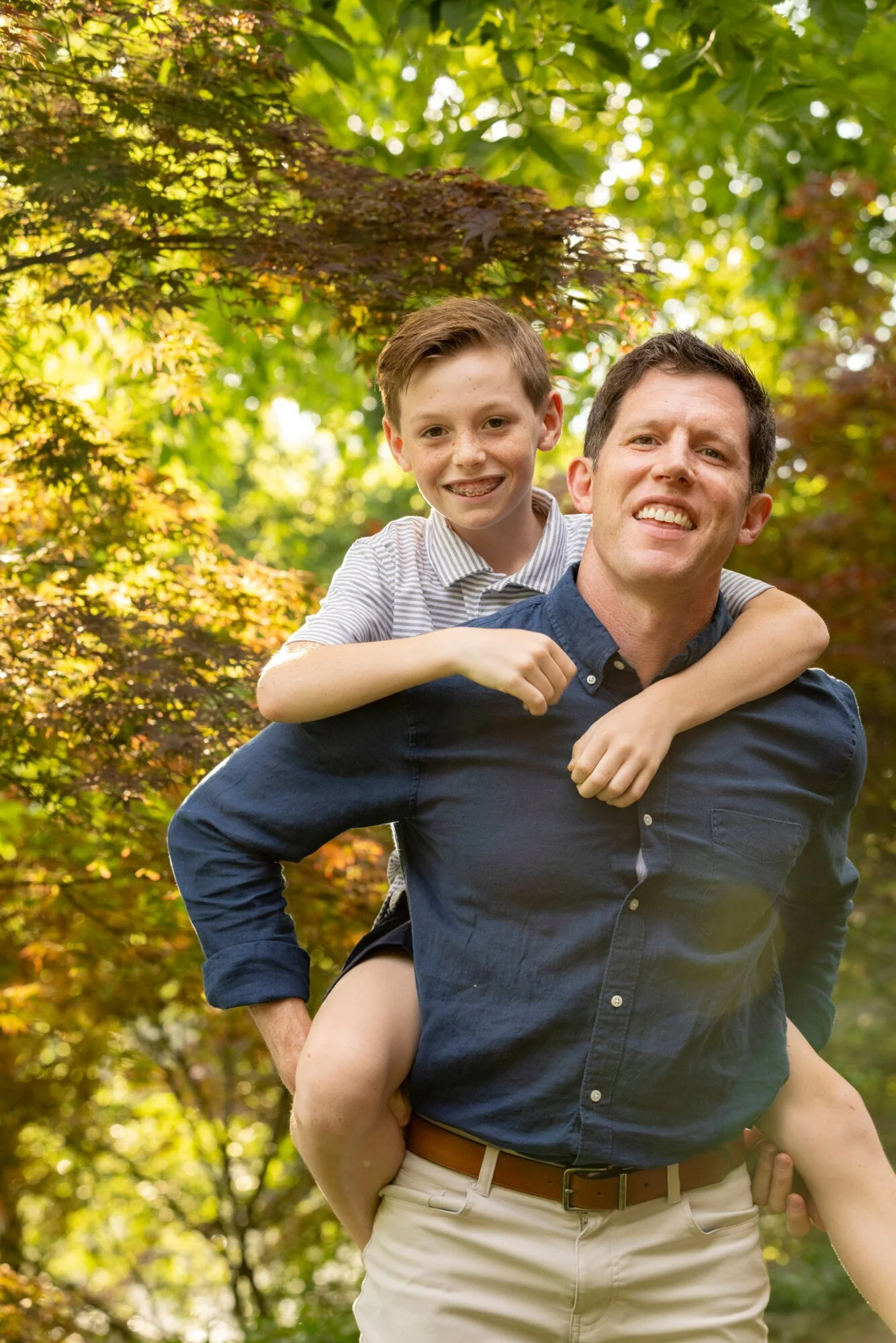 A man wearing a dark blue shirt carrying a boy on his shoulders in a park with green and fall-colored leaves in the background.