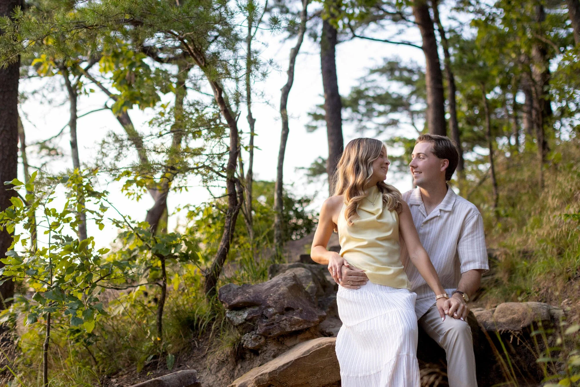 A young woman and a young man sitting on rocks in a wooded area. The woman is wearing a cream-colored top and a white skirt, while the man is wearing a striped shirt. They are smiling at each other and seem to be enjoying the outdoor scenery surround