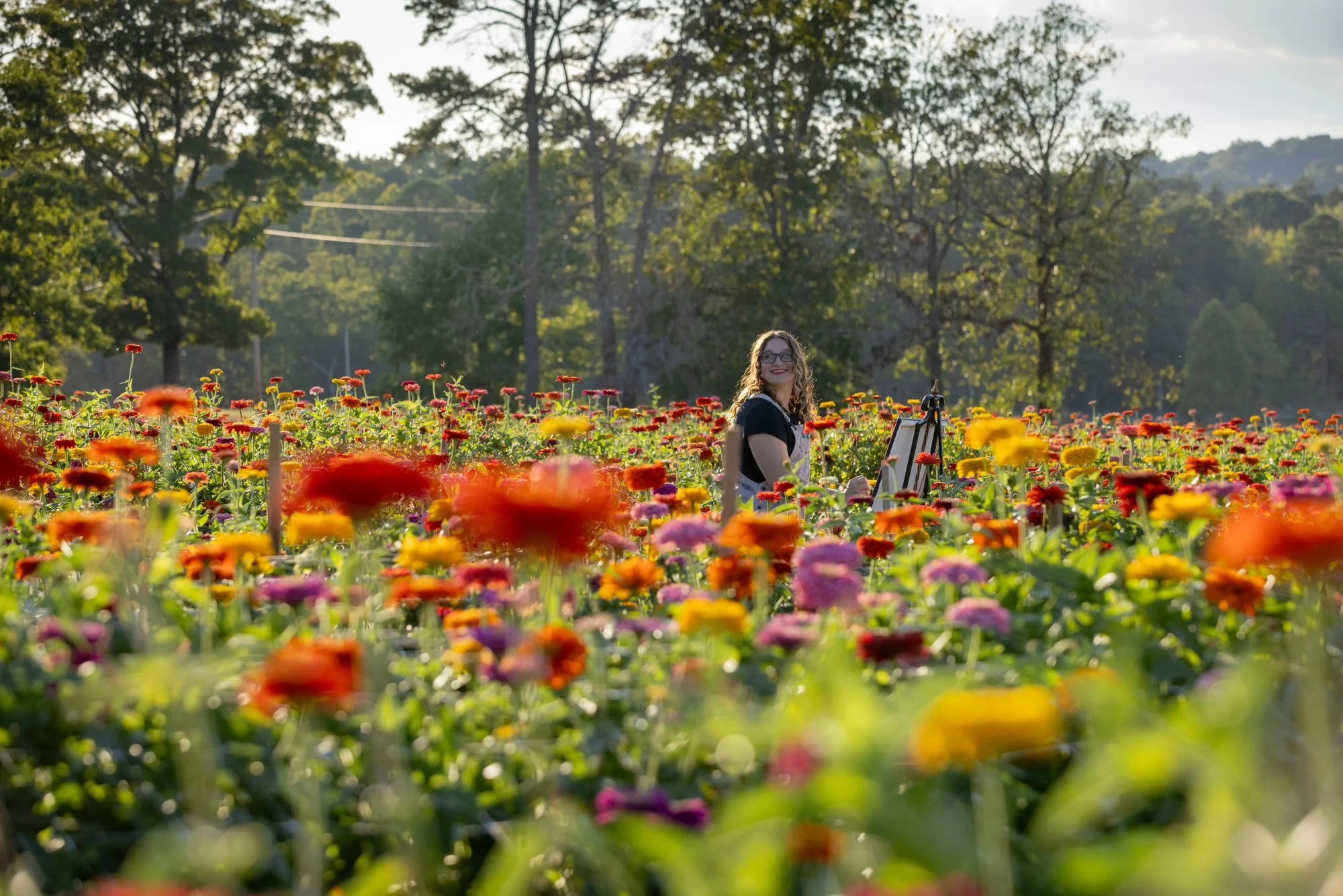 A woman with curly hair and glasses smiling while painting in a colorful flower field with trees and hills in the background.