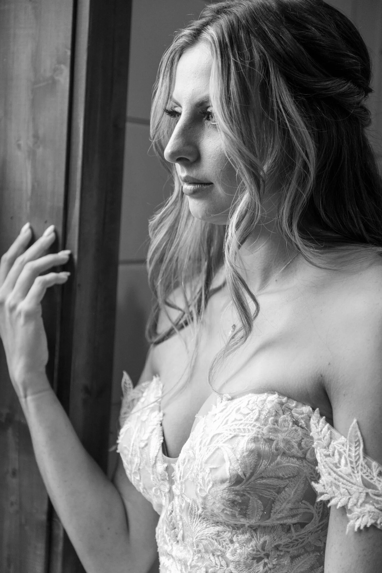 A black-and-white photo of a woman with long, wavy hair looking down, wearing an off-shoulder lace dress, touching a wooden door frame with her left hand.