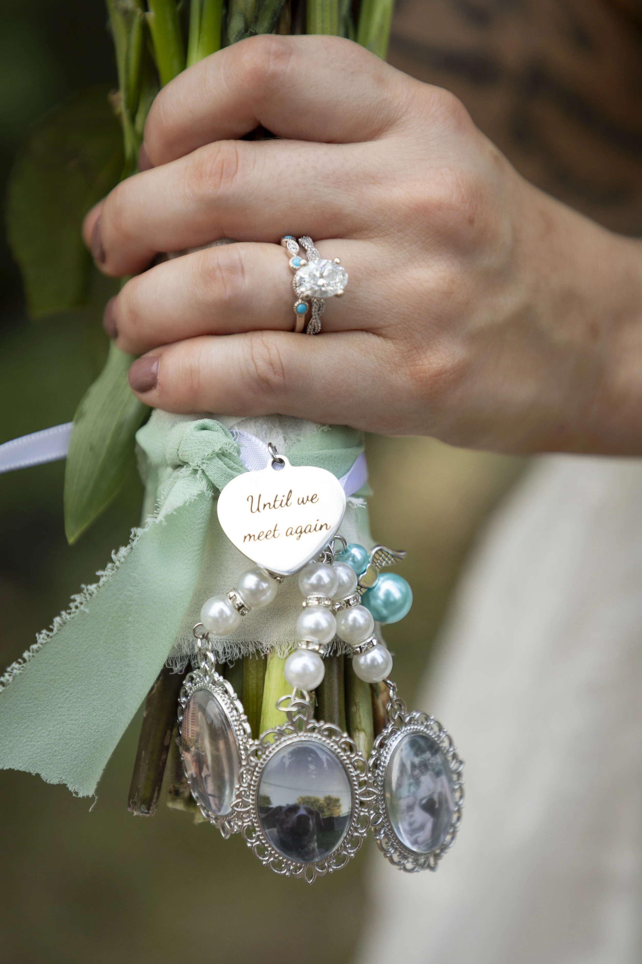 Close-up of a person's hand holding a bouquet of flowers, with a large diamond ring on their finger and several pieces of jewelry hanging from the bouquet, including a small heart-shaped charm with the words "Until we meet again."