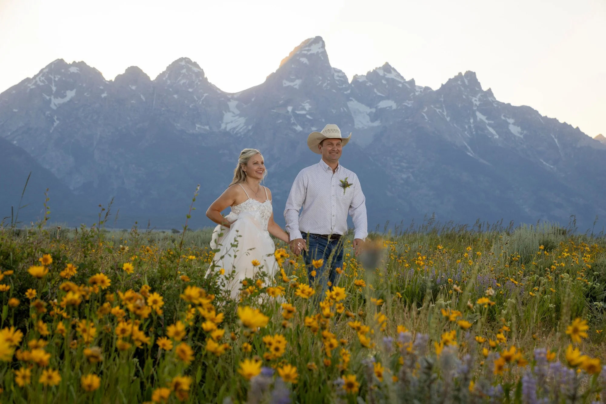 A smiling couple holding hands in a yellow wildflower field with tall mountains in the background.