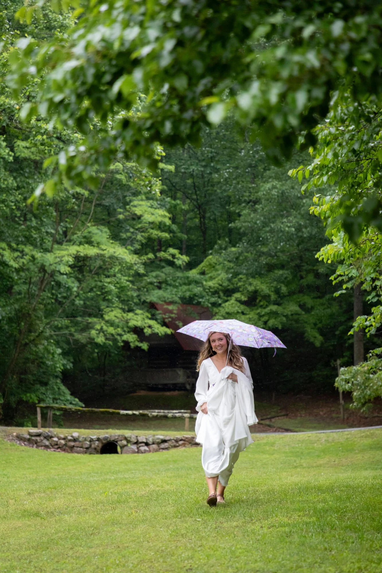 A woman in a white dress holding a purple floral umbrella walking on a grassy lawn with trees and a small bridge in the background.