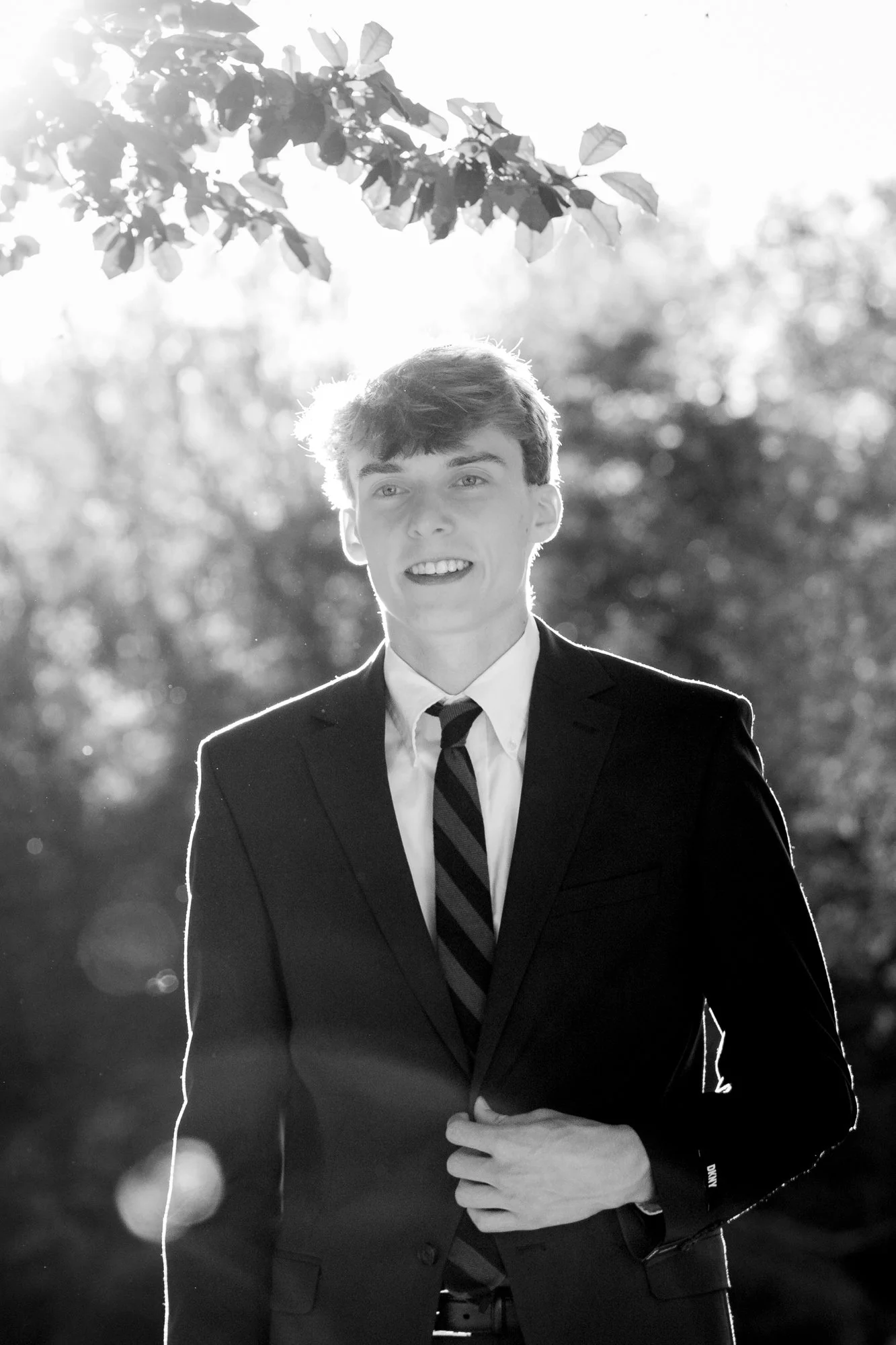 A young man in a suit and tie smiling outdoors with sunlight behind him, standing near tree branches and leaves in black and white.