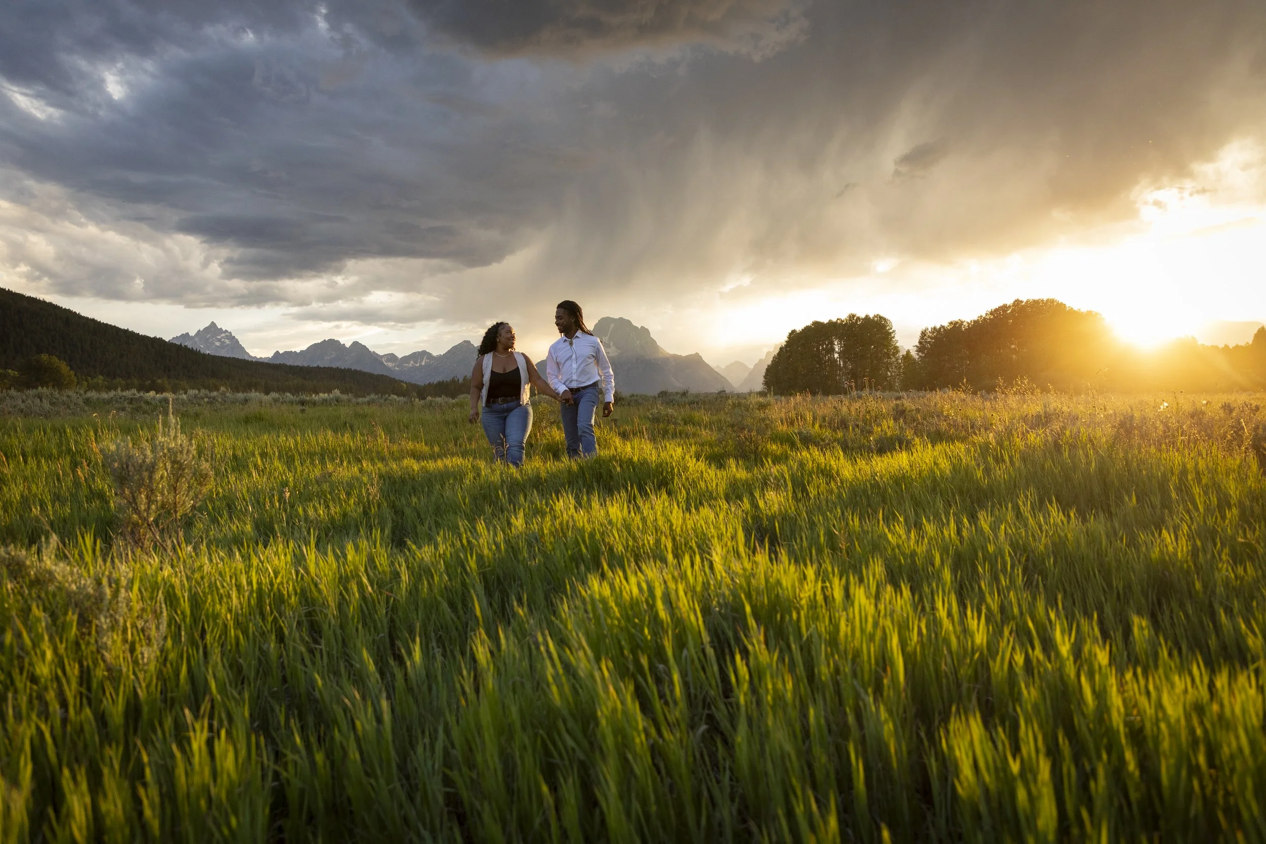 A couple walking hand in hand through a grassy field at sunset with mountains in the background and dark clouds overhead.