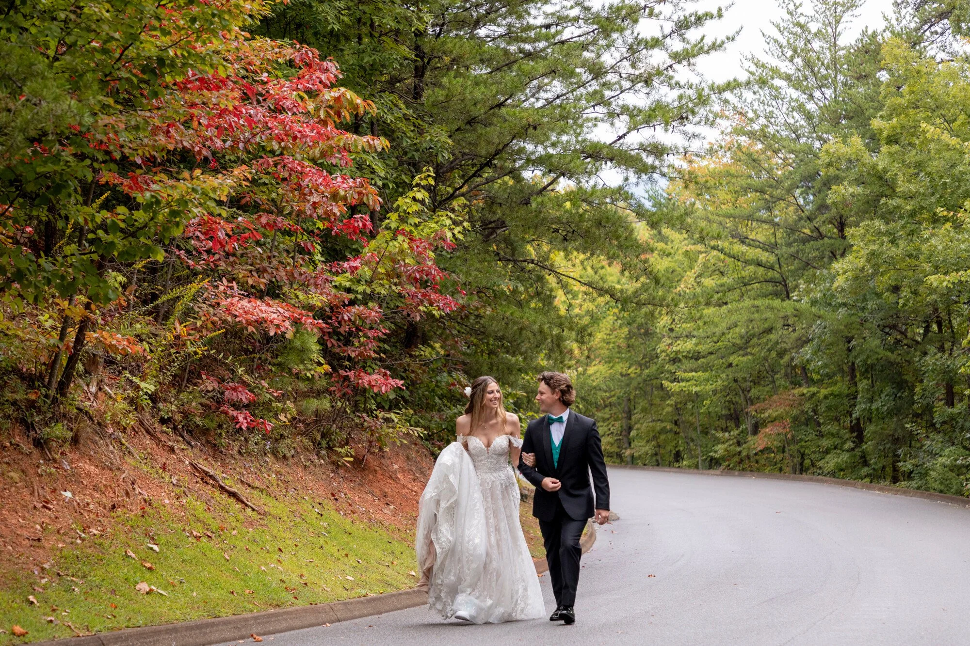 A bride and groom walking hand in hand on a sidewalk beside a park road, surrounded by lush green trees and some red and yellow fall foliage, during daytime.